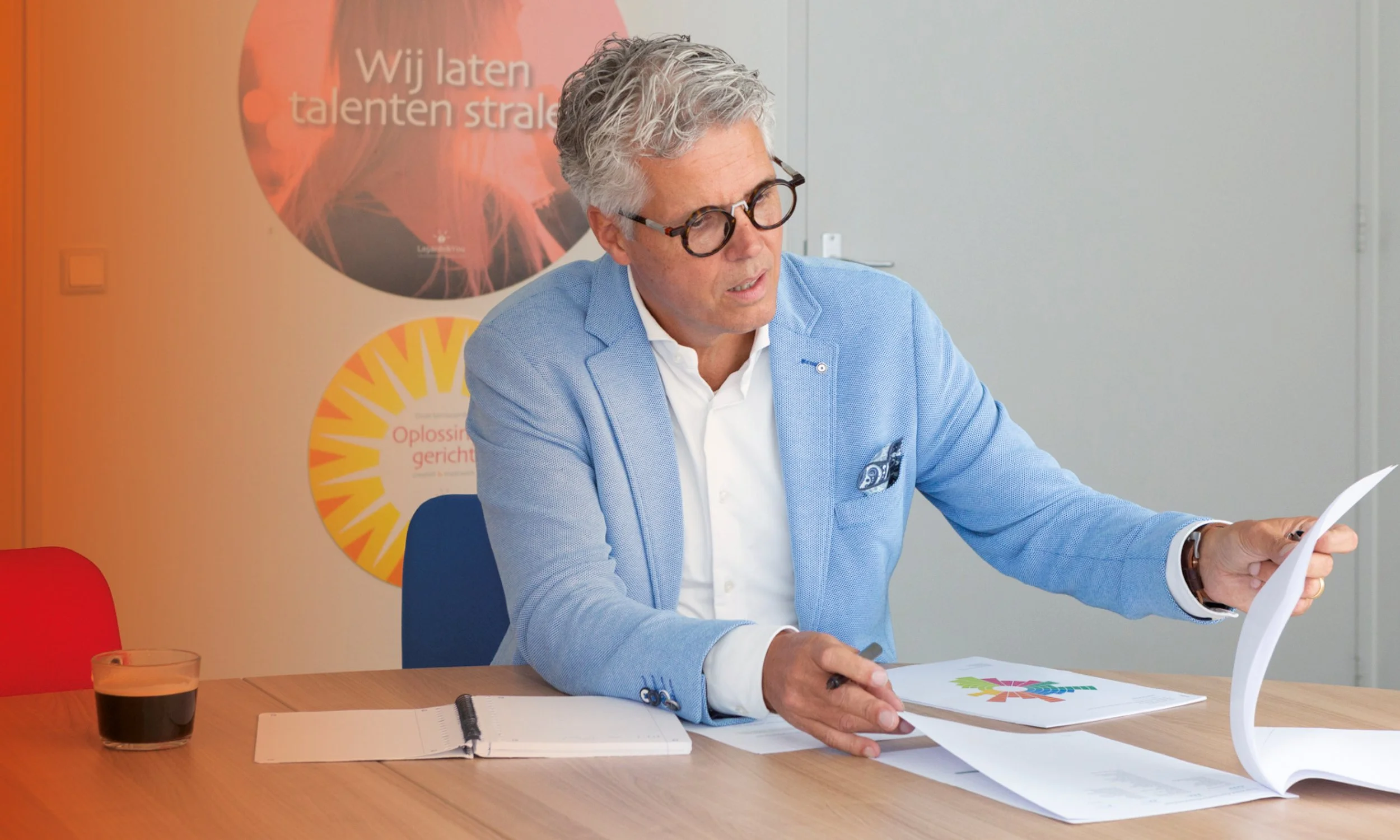 Een man met grijs haar, bril en een lichte blauwe jas zit aan een tafel met documenten, een glas cola en een notitieboekje en spreekt of presenteert tijdens een zakelijke bijeenkomst.