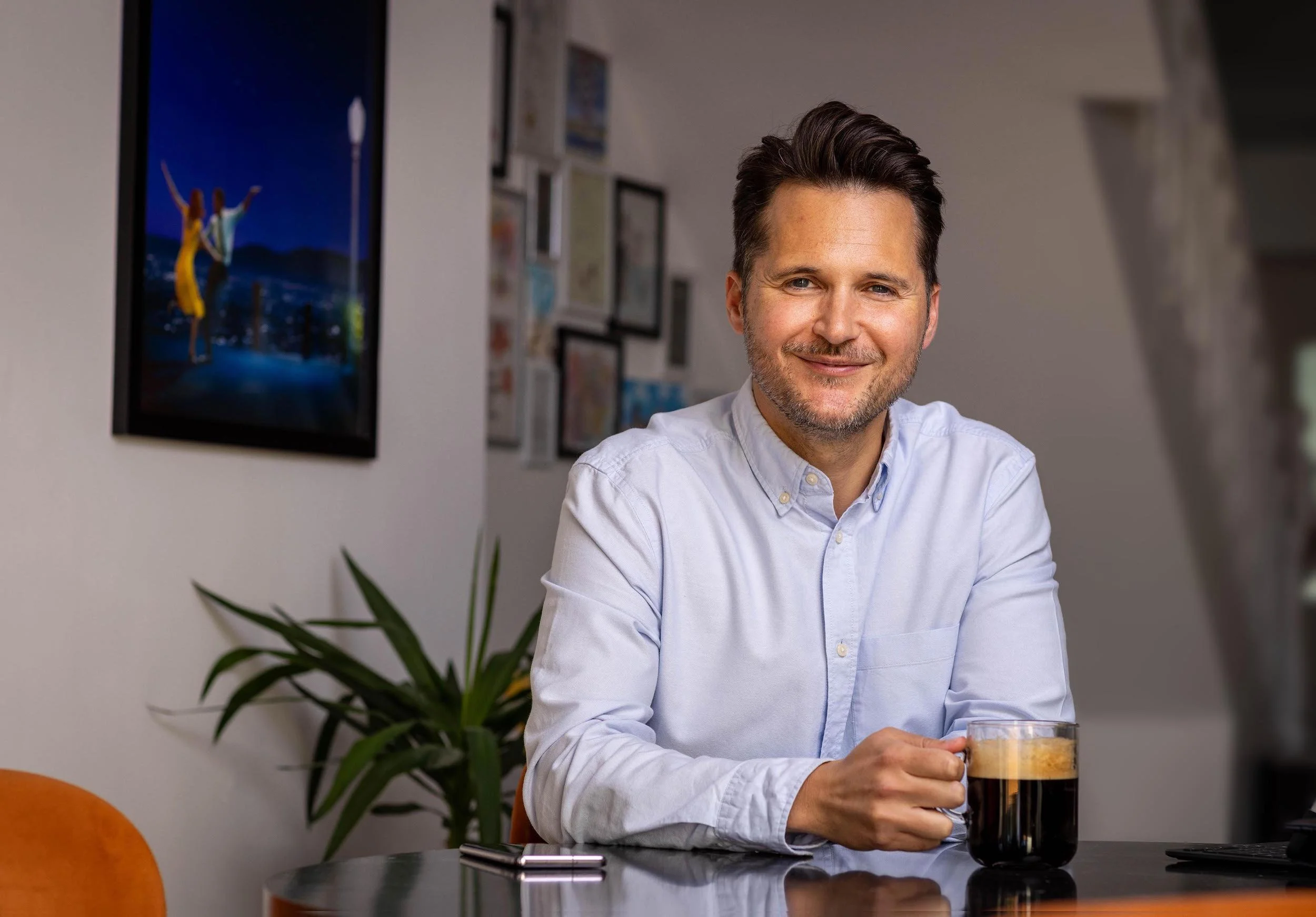 Man sitting at a table with a cup of coffee, holding the handle and smiling. Background includes a framed wall art and indoor plant.