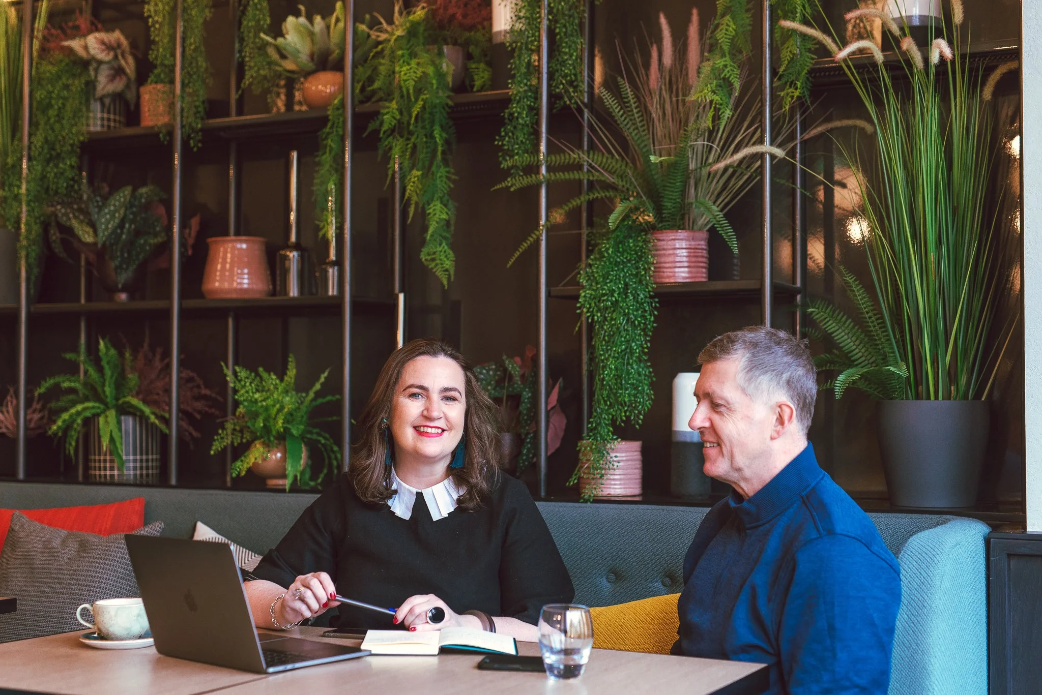 Webdesigner Lucy O'Reilly sitting at a table in a cozy cafe with a client, using a laptop and a notebook.