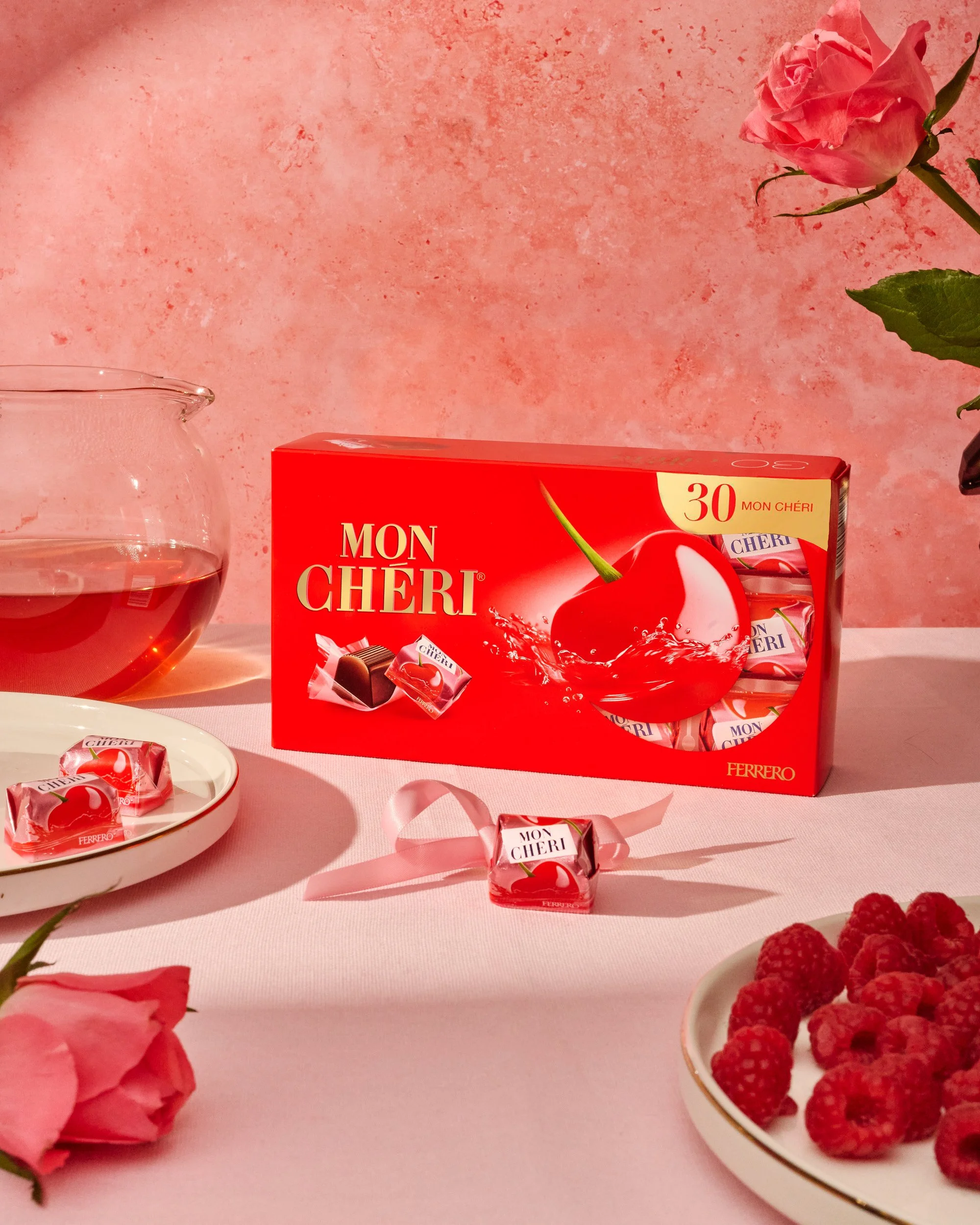 Boîte de chocolats Mon Chéri de Ferrero à la cerise avec un verre d'eau, une assiette de framboises, une rose rose et un fond rose.