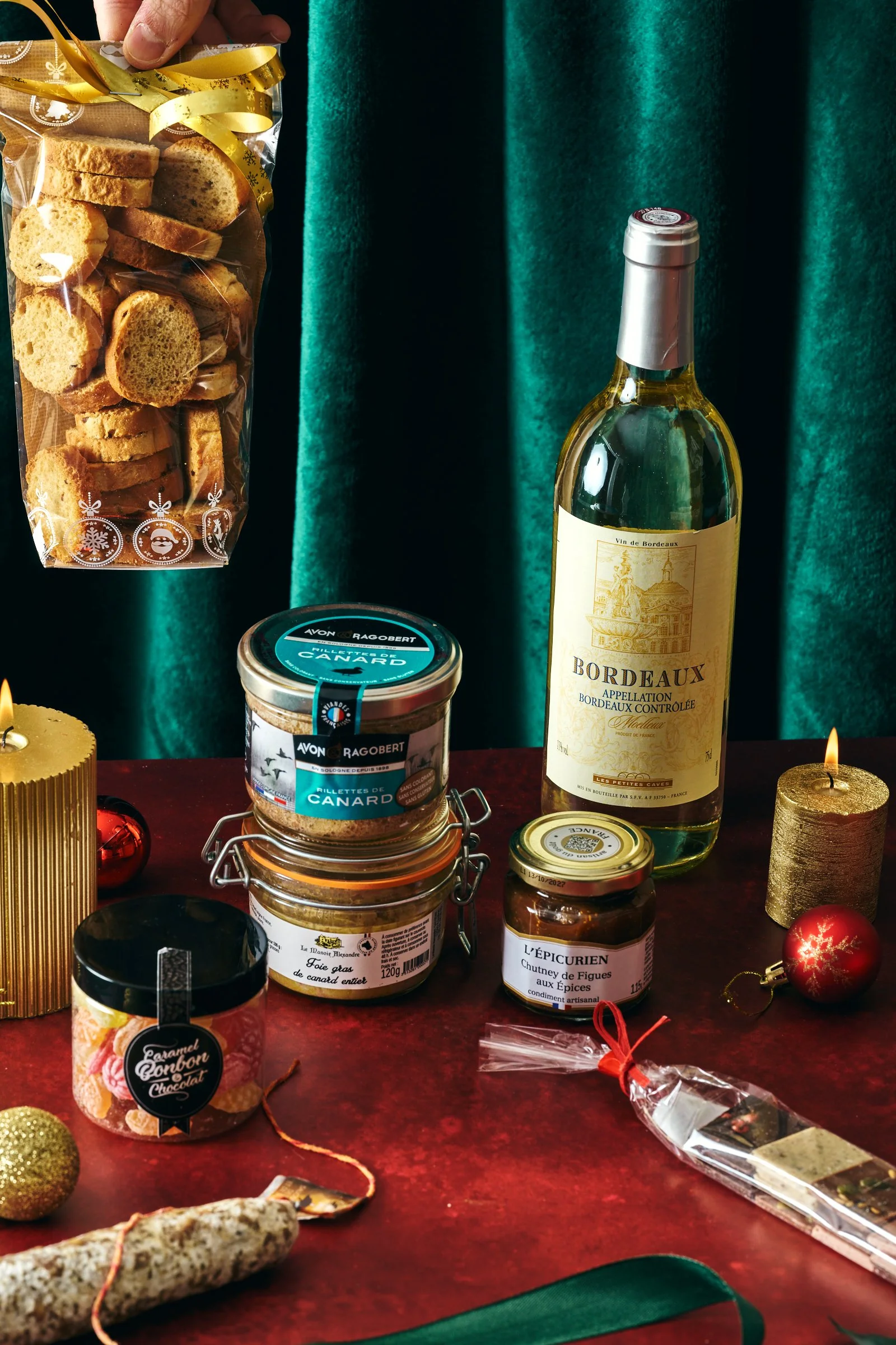 Une bouteille de vin blanc, des bocaux de foie gras, du chutney de figues, des biscuits, une bûche de Noël, des bougies, et des décorations de Noël sur une table en bois.