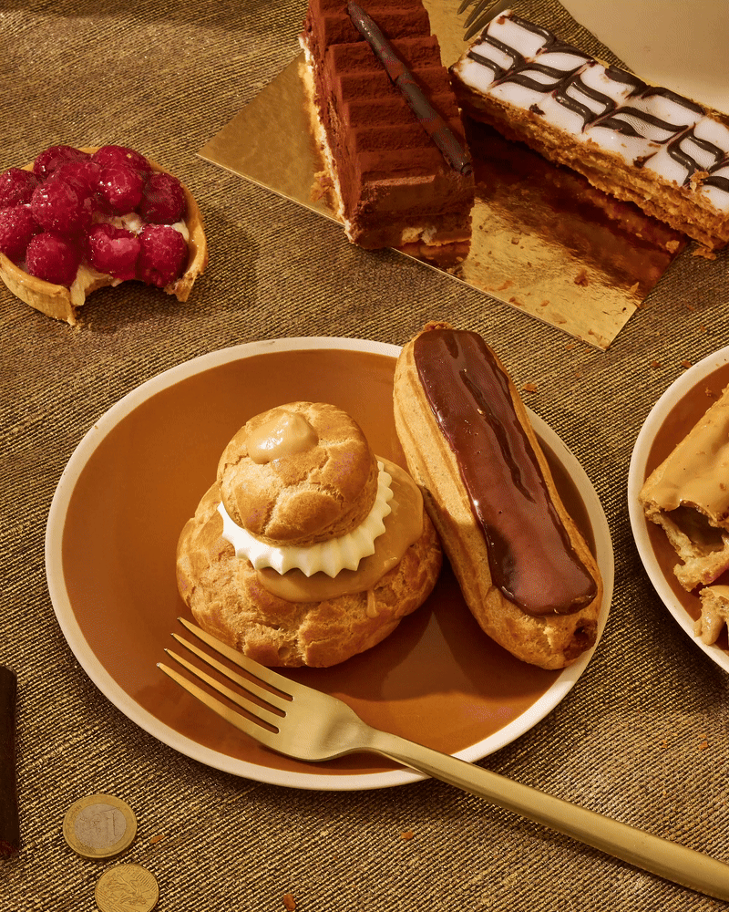 Assiette avec une profiterole garnie de crème, un éclaire au chocolat, et une tarte aux framboises avec une part manquante. En arrière-plan, deux gâteaux chocolat et chantilly.