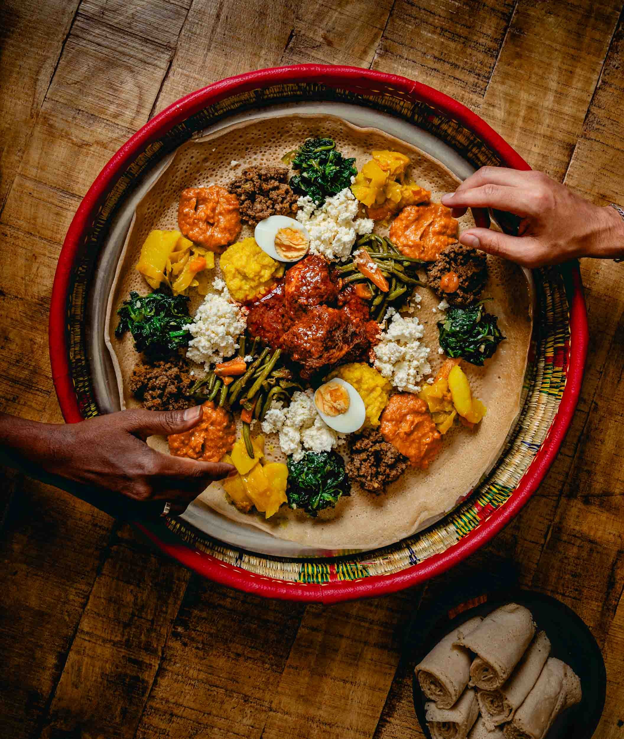 Un plateau d'injera éthiopien avec divers plats, y compris des légumes, des sauces, et des œufs, entouré de mains attrapant la nourriture.