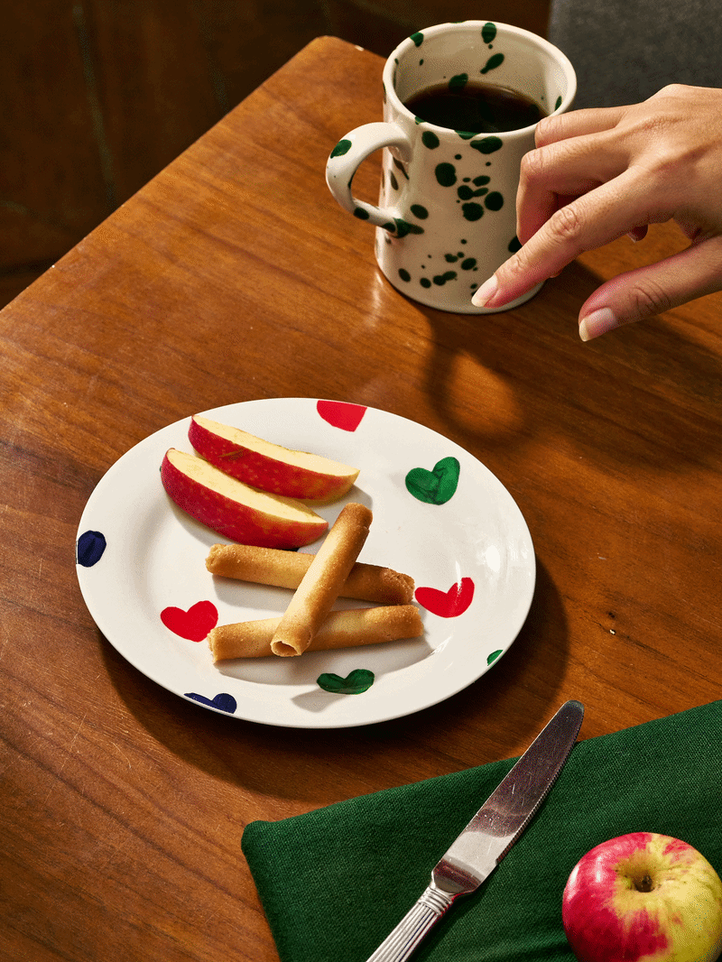 Une tasse de café ou de thé noir avec une main qui la tient, une assiette blanche avec des tranches de pomme rouge et des rouleaux de pâtisserie, une fourchette sur une serviette verte, et une pomme rouge sur la table en bois.