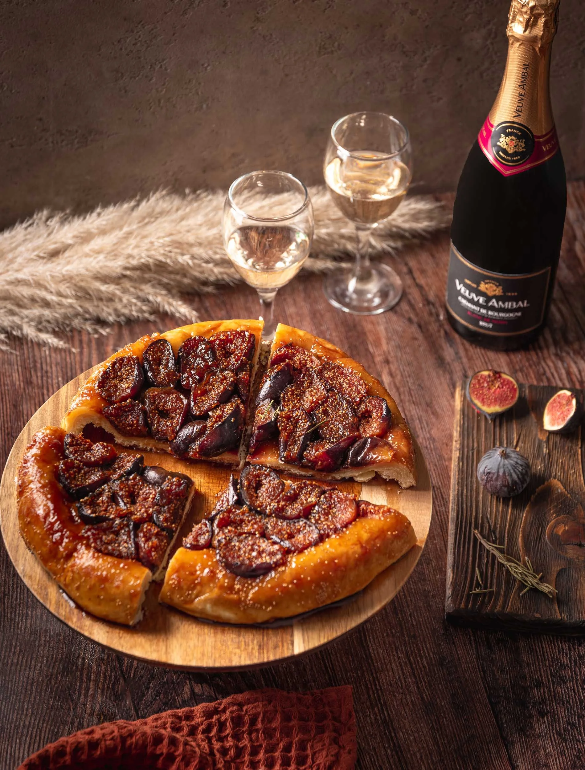 Une tarte aux figues sur une planche en bois, deux verres de vin blanc pétillant et une bouteille de champagne Veuve Ambal posés sur une table en bois avec des éléments de décoration rustiques.