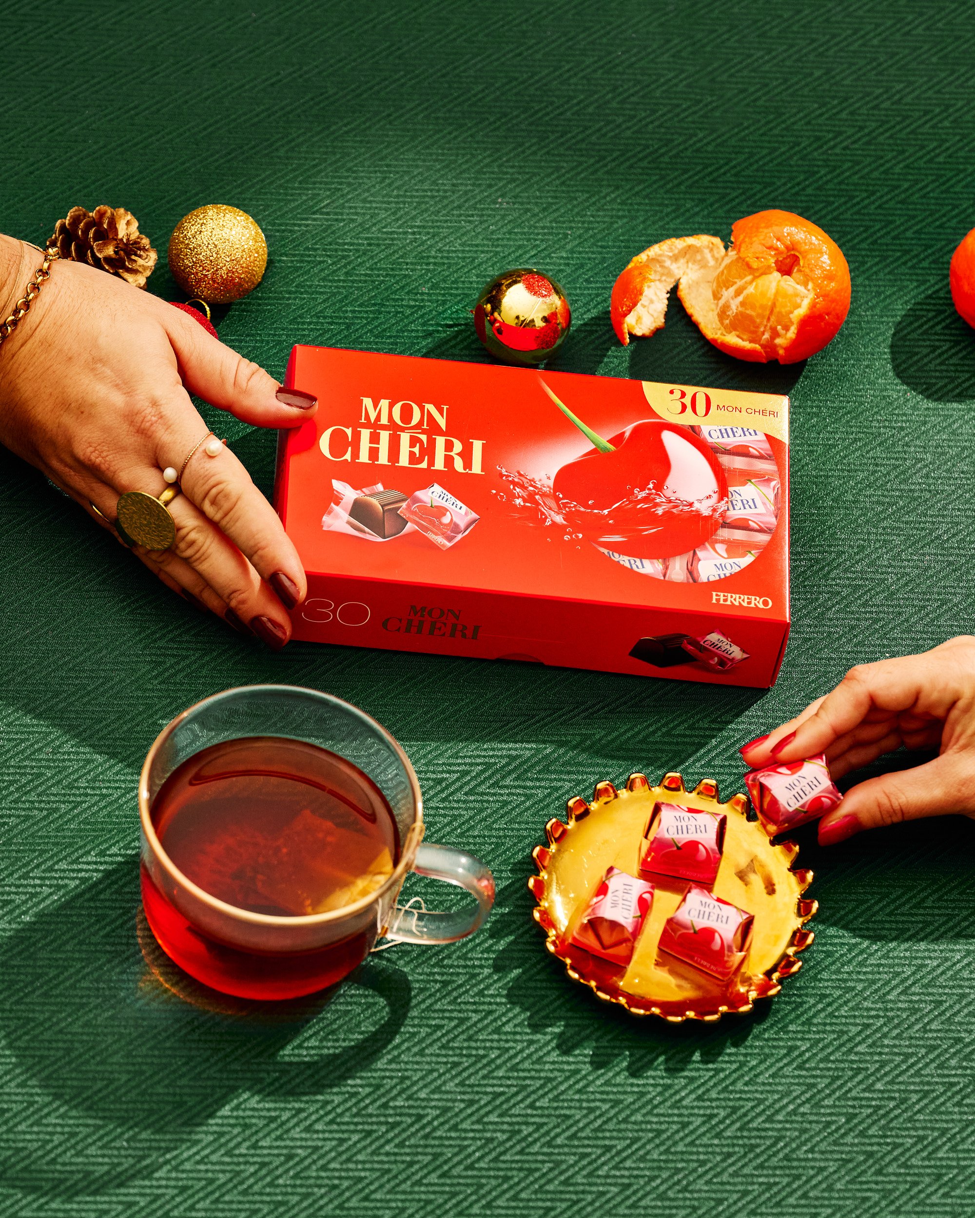 Une boîte de chocolat Mon Chéri de Ferrero est posée sur une table verte, entourée de décorations de Noël, d'une tasse de thé et d'une coupelle contenant des chocolats discutés.