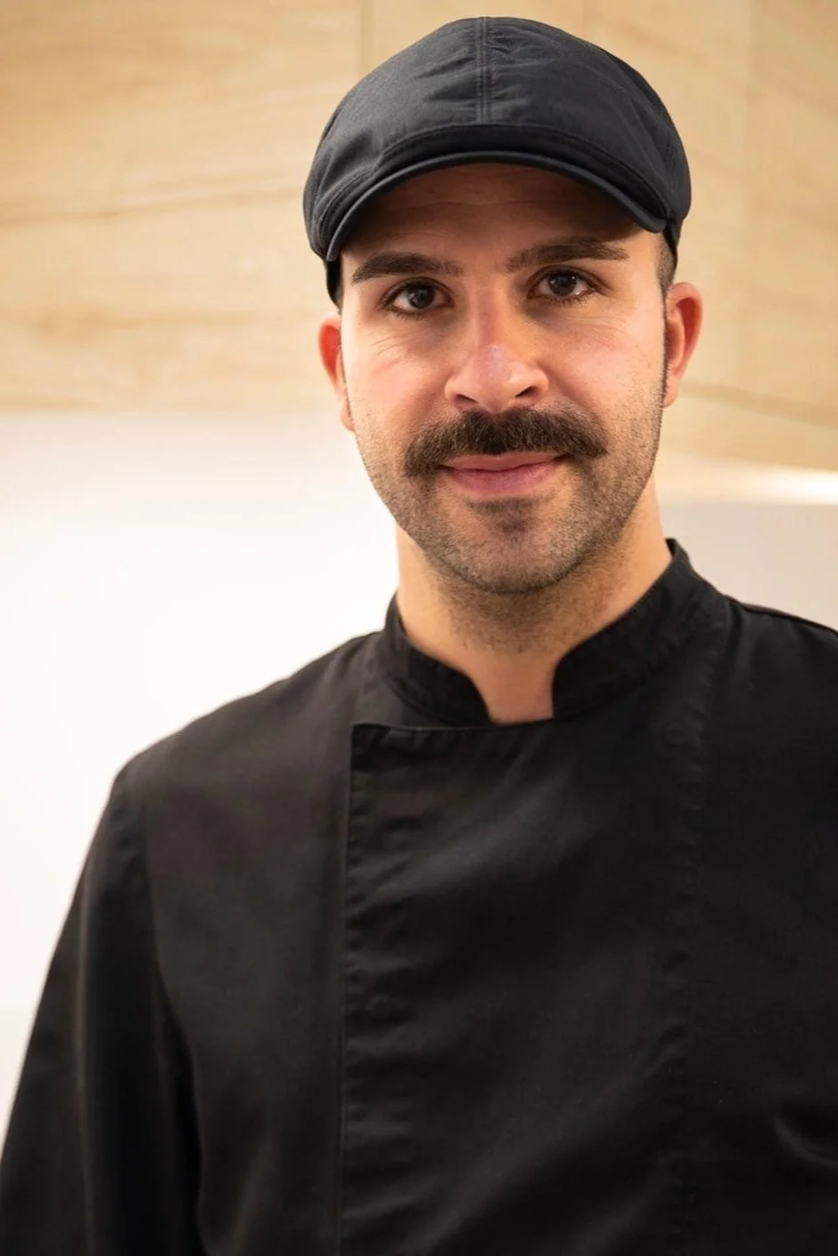 Homme moustachu portant une casquette et une veste noire debout devant un arrière-plan neutre.