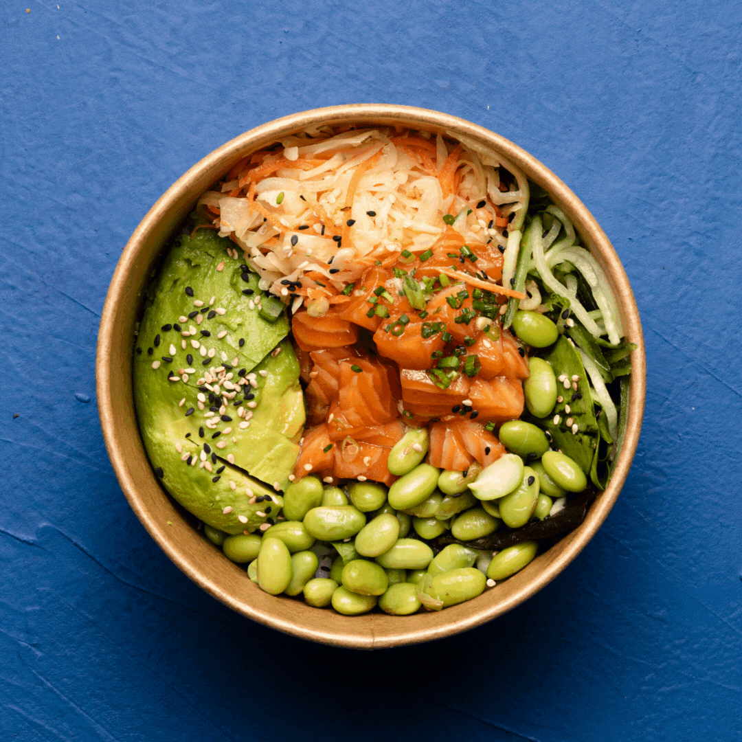 Bol de poké avec saumon, avocat, edamame, choux râpé, concombre, graines de sésame, et riz, sur fond bleu.