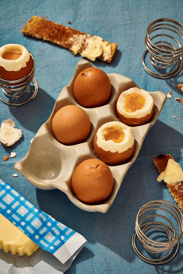 Oeufs à la coque dans un coquetier, avec des tartines beurrées sur une nappe bleue.