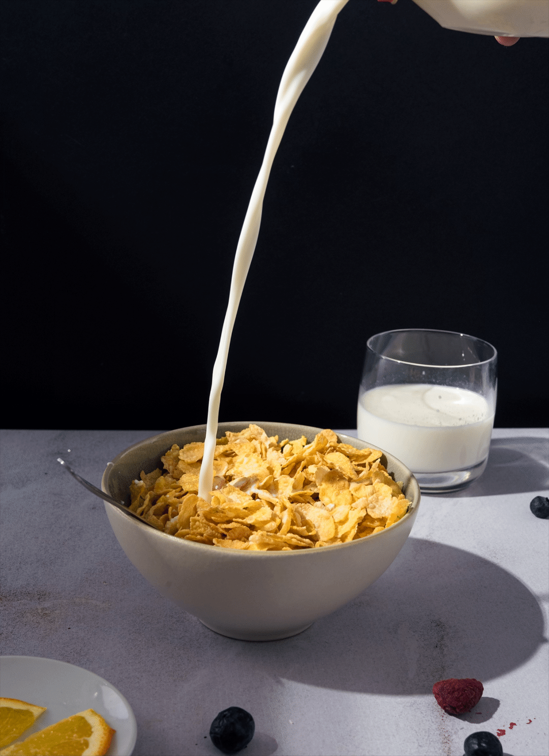 Un bol de céréales avec du lait versé dedans, accompagné d'un verre de lait à côté. Il y a aussi des morceaux d'orange et des baies autour.