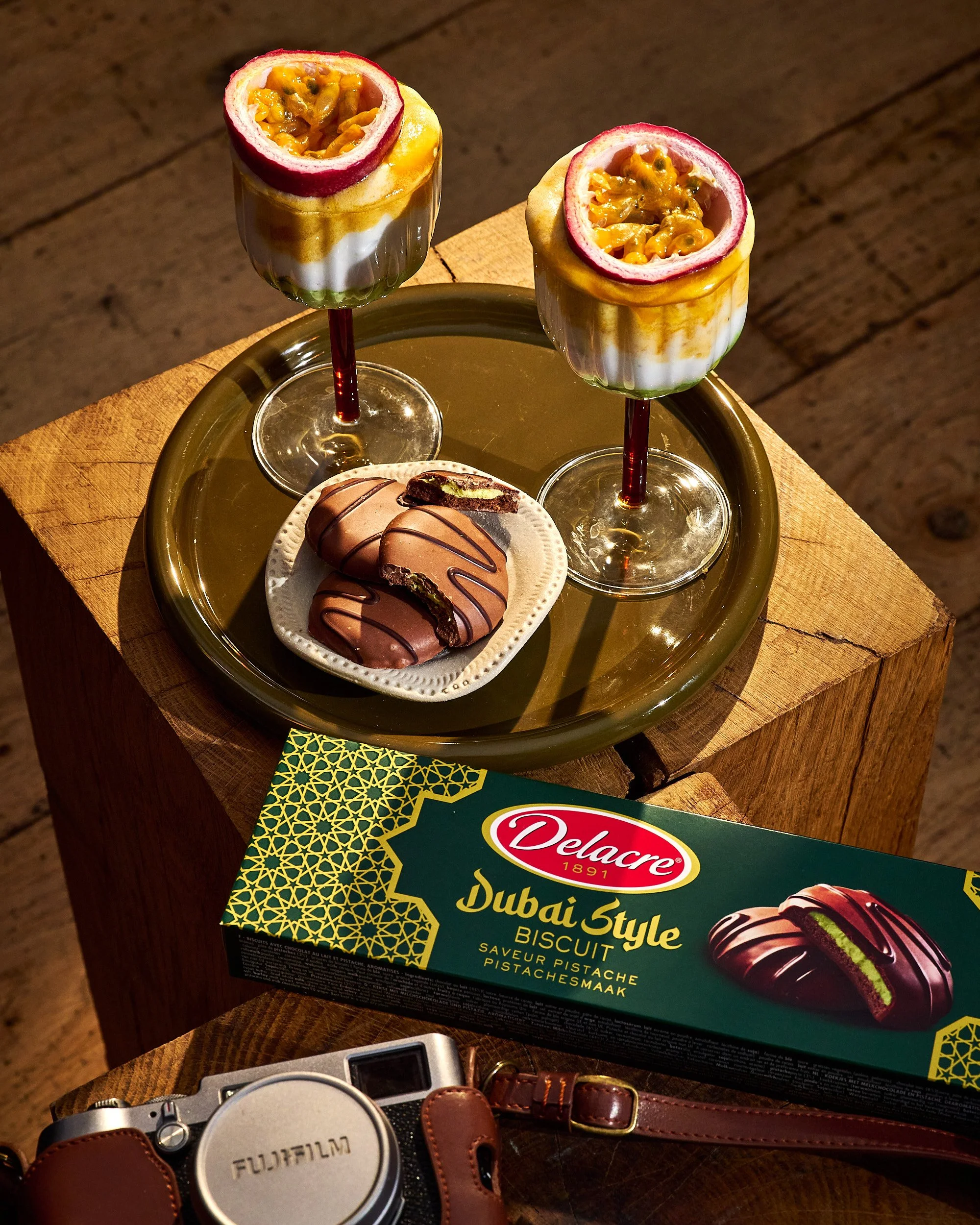 Deux coupes de dessert avec passion fruit coupé en haut, quelques biscuits chocolatés accompagnés, une boîte de biscuits Delacre Dubai Style Biscuit pistache. On voit également un appareil photo Fujifilm et un sac en cuir sur une table en bois. Photo