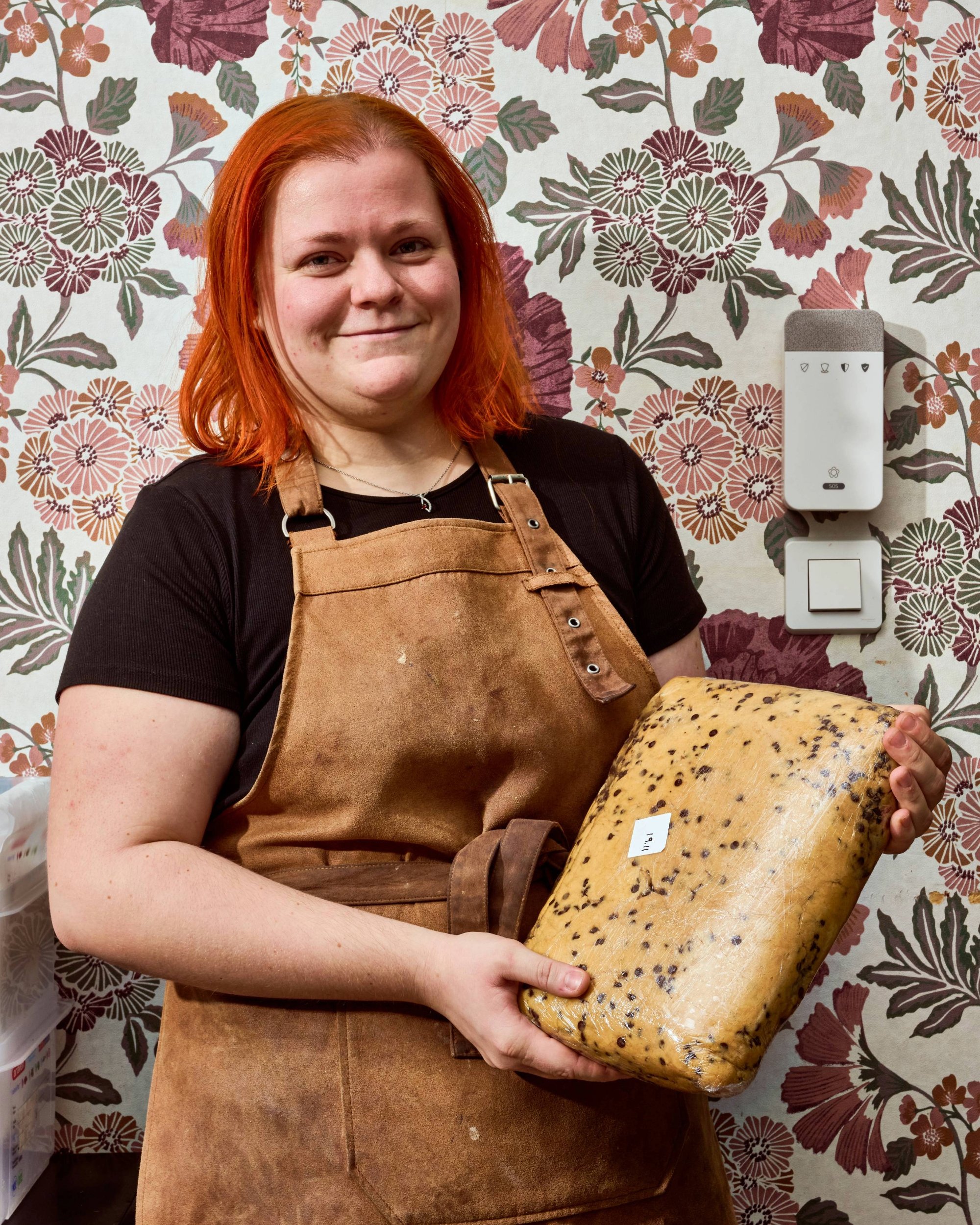 Personne souriante avec des cheveux roux tenant un gros morceau de fromage avec des grains de poivre, portant un tablier marron. Fond avec motif floral.