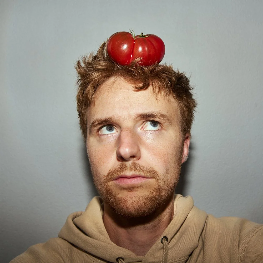 Un homme avec des cheveux bruns et une barbe, portant un sweat beige, regarde vers le haut avec deux tomates rouges sur sa tête.