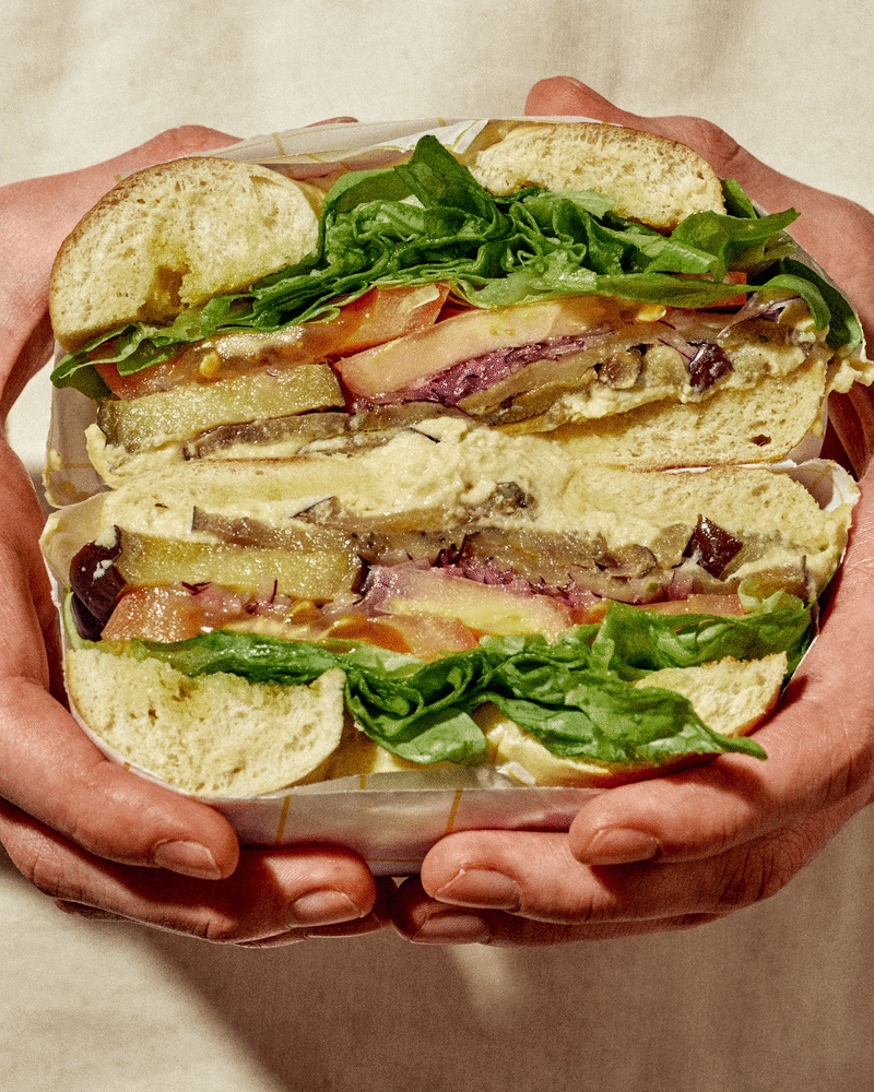 Sandwich coupé en deux avec de la laitue, tomate, viande, fromage, et champignons, tenu par des mains.  Pris en photo par le photographe culinaire Nicolas Blum