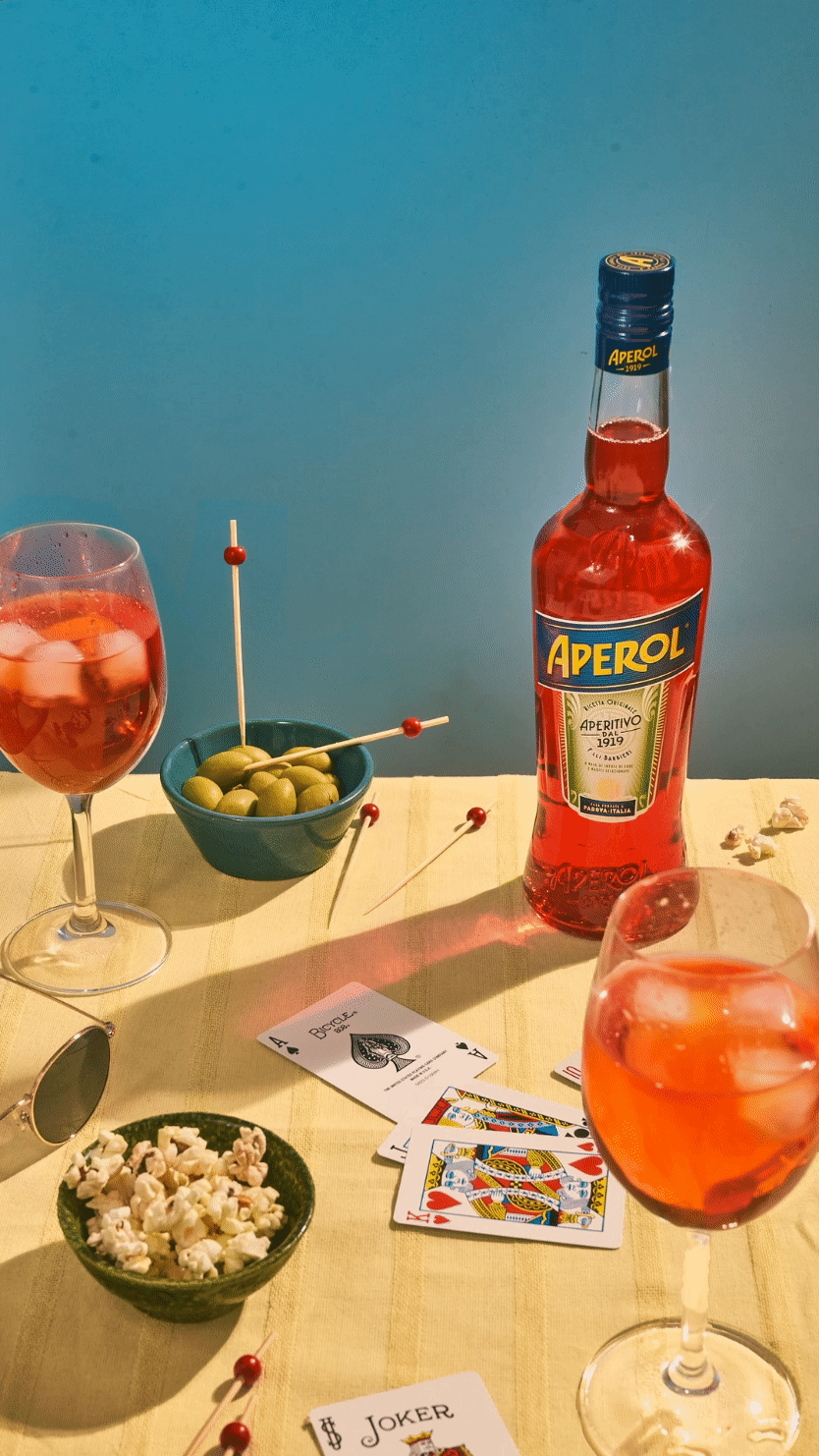 Bouteille d'Aperol, verres de cocktail Spritz, olives, popcorn, lunettes de soleil, cartes à jouer sur une table.