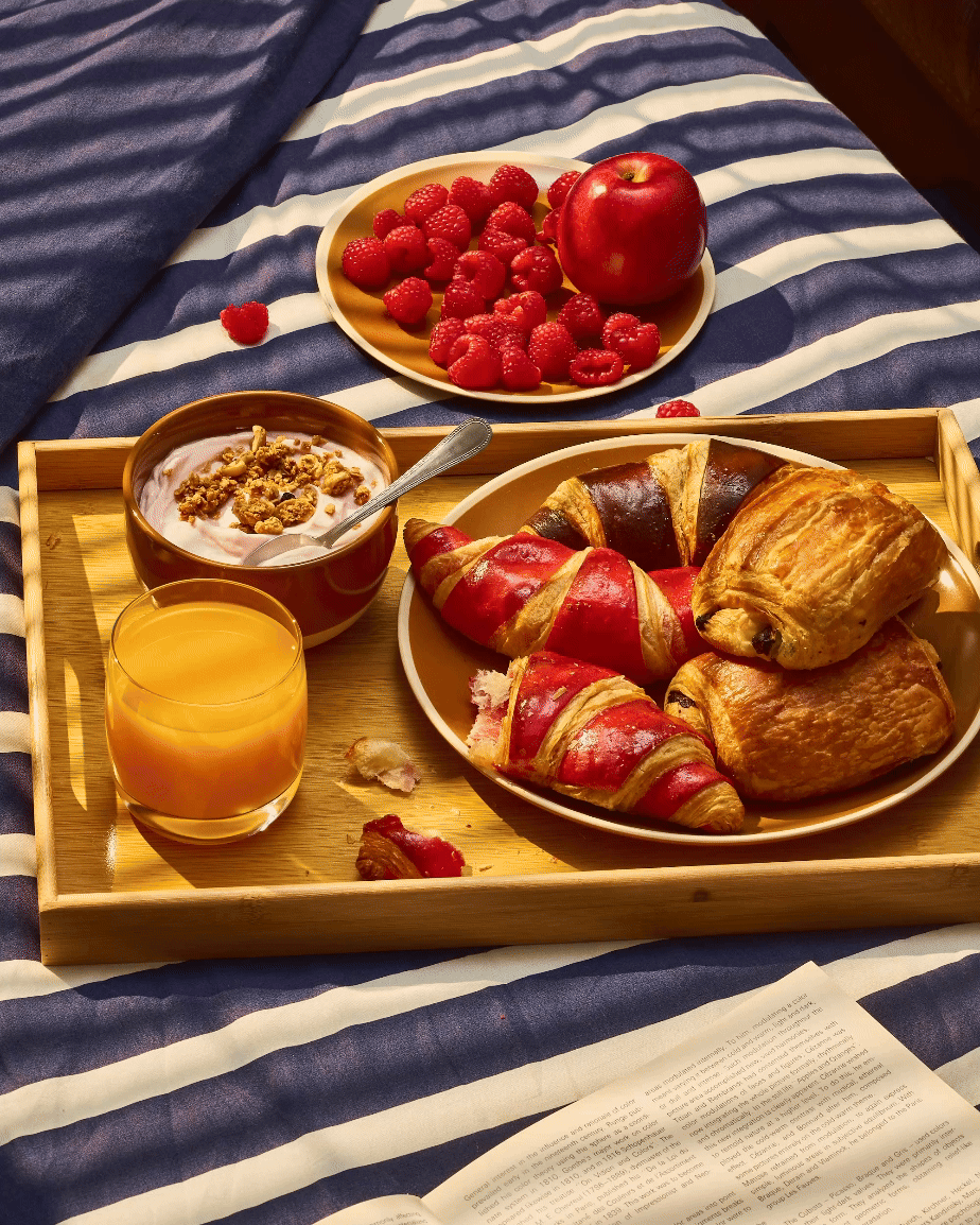 Petit déjeuner avec croissants et chocolatines, yaourt au granola et verre de jus d'orange sur un plateau, framboises et pomme sur une assiette. Livre ouvert sur des draps rayés bleus et blancs.