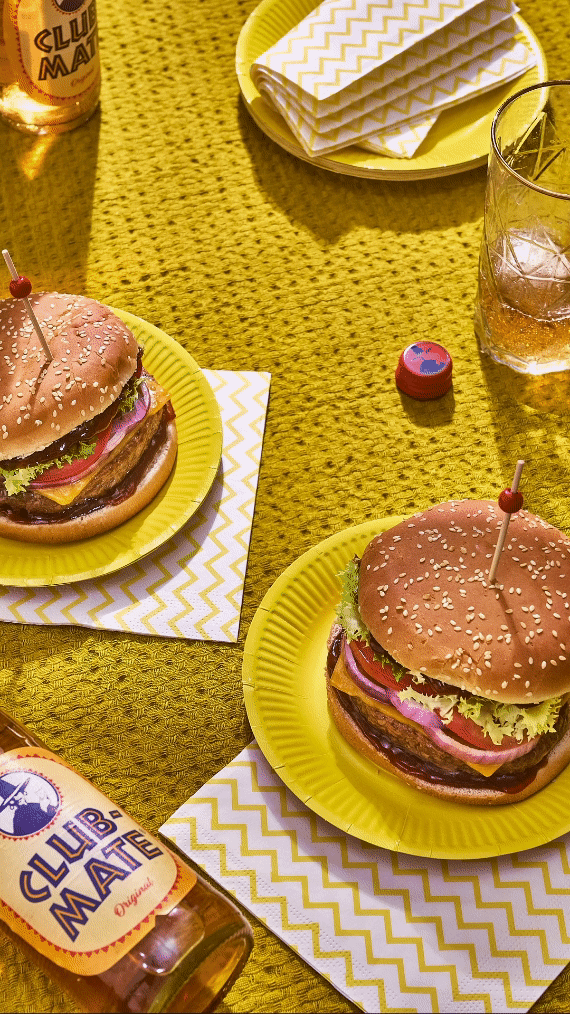 Deux hamburgers sur des assiettes jaunes, avec des serviettes à motifs zigzag jaunes et blancs, et des bouteilles de Club-Mate sur une couverture jaune.
