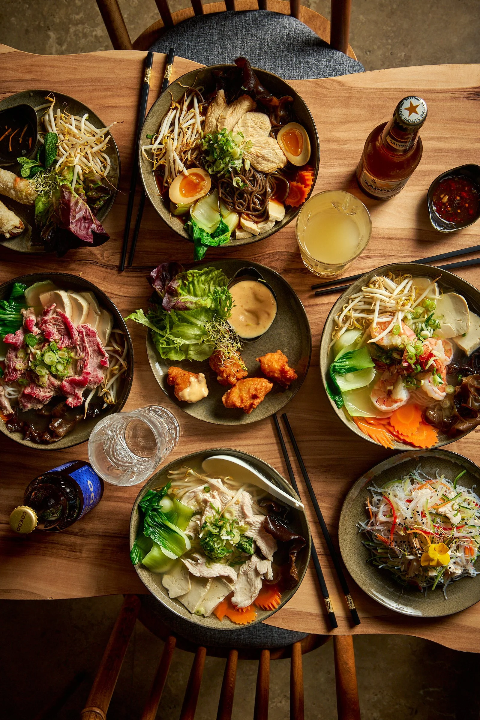 Table avec plusieurs plats de cuisine asiatique, notamment des ramen, des salades et des garnitures, ainsi que des boissons comme une bière, un soda et un verre de citronnade.