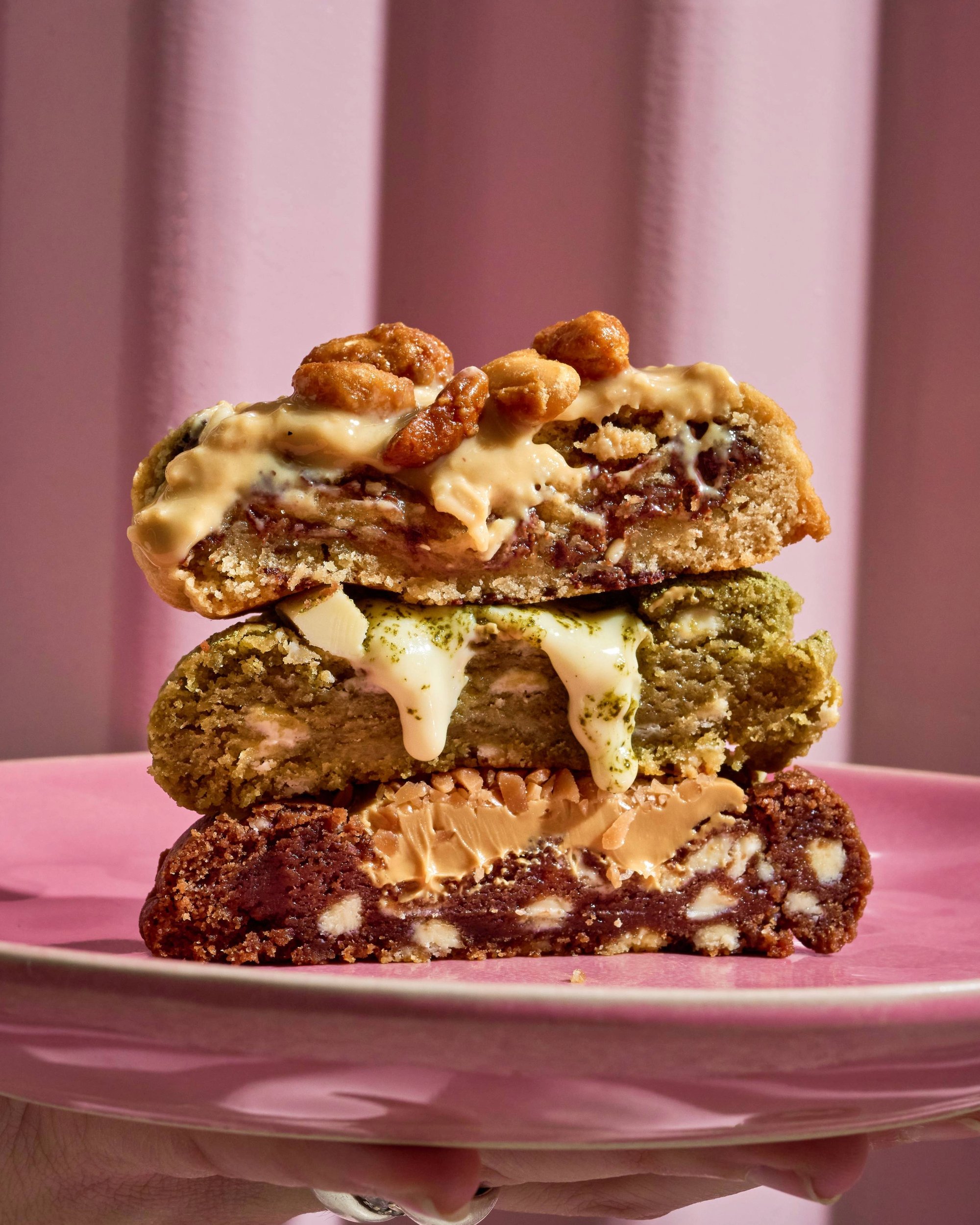 Trois cookies empilés avec garnitures variées sur une assiette rose, devant un fond rose.