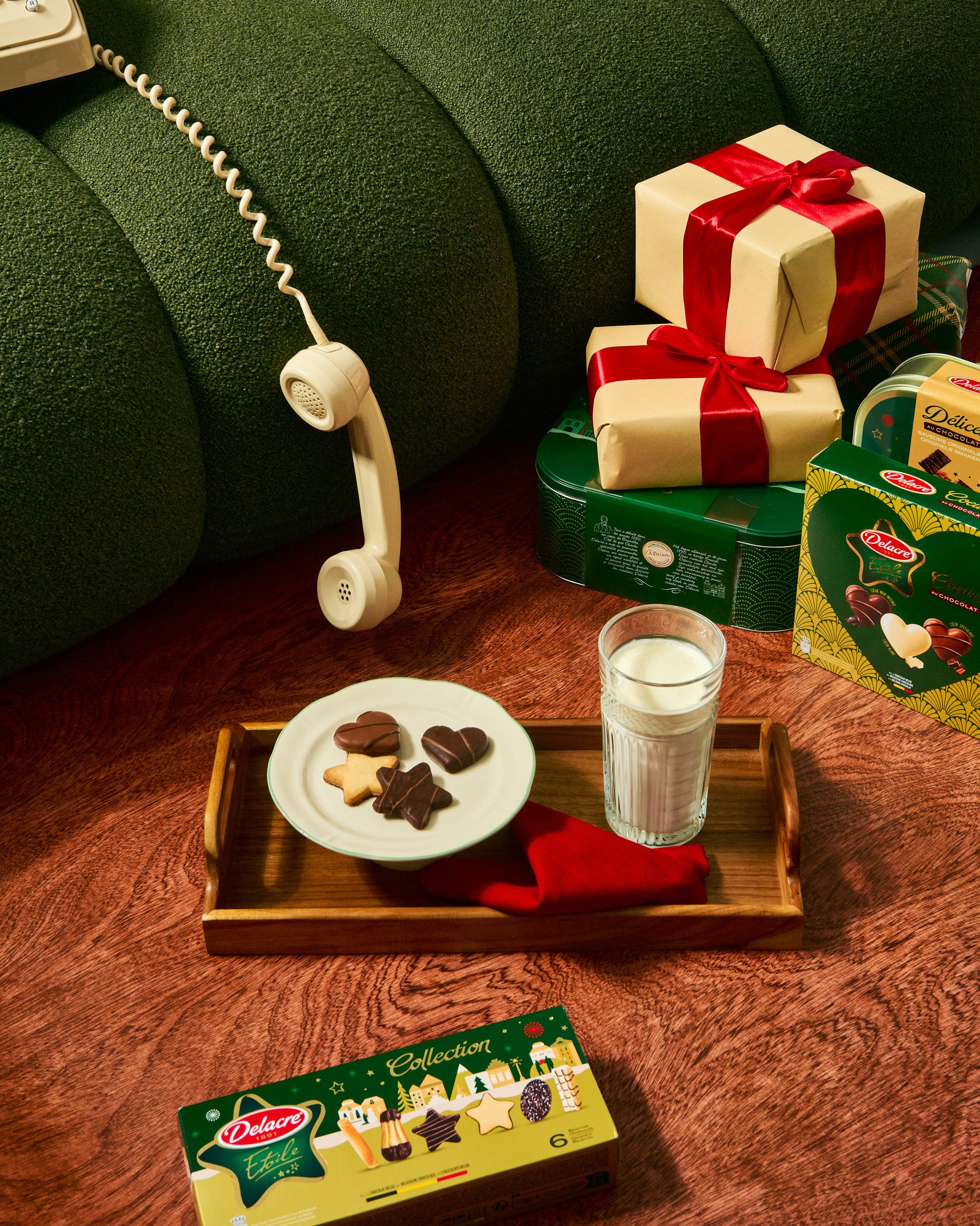 Une table avec des biscuits en forme de cœur et d'étoile, un verre de lait, plusieurs boîtes de chocolats, un plateau en bois avec un napperon rouge et un téléphone vintage suspendu à un canapé vert.