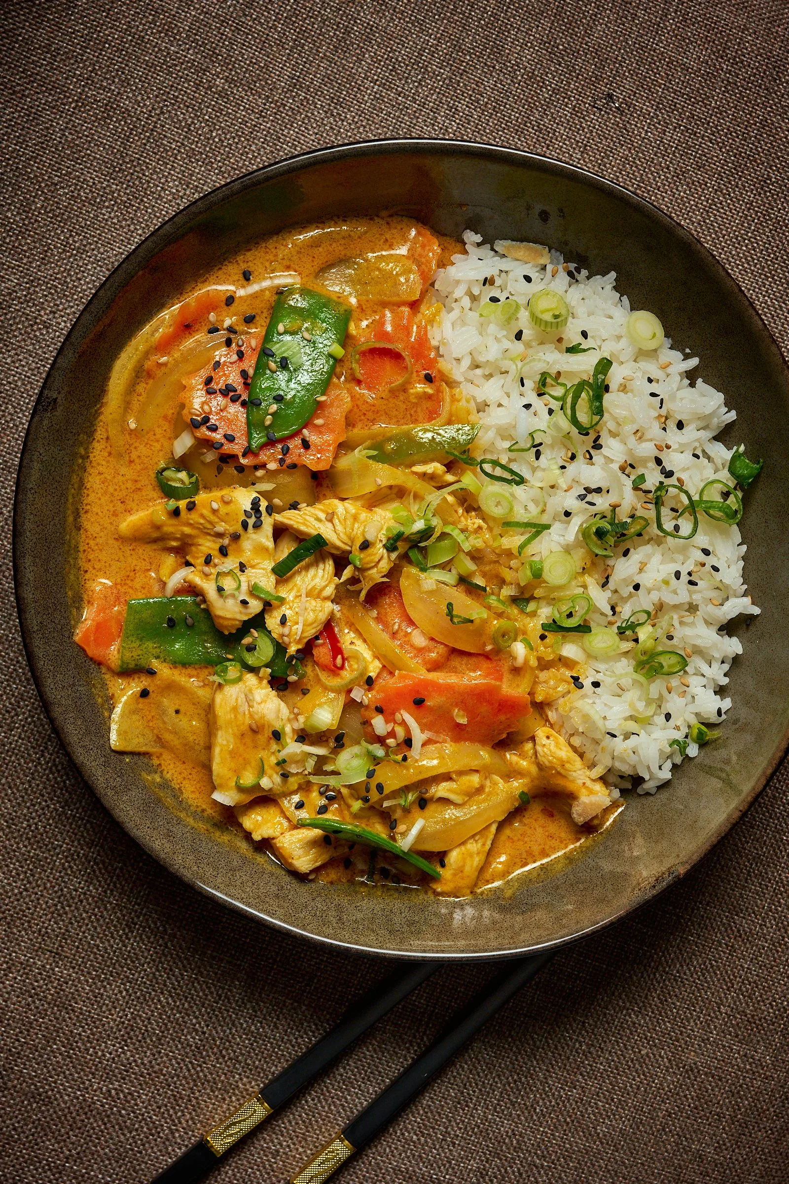 Un plat de riz blanc et un curry thaï avec poulet, légumes comme poivrons, tomates, et oignons, garnis de graines de sésame noires et de ciboulette, présenté dans une assiette noire sur une nappe en tissu.