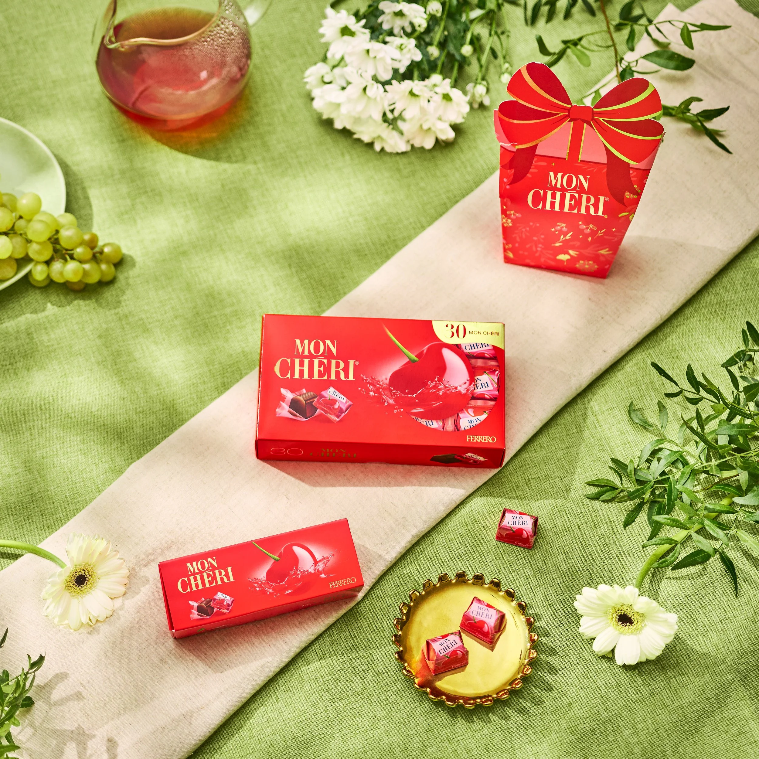 Gamme de chocolats Mon Chéri de Ferrero sur une nappe verte avec des fleurs blanches et un panier en or, accompagnée d'une bouteille de vin rosé et de grappes de raisins verts.
