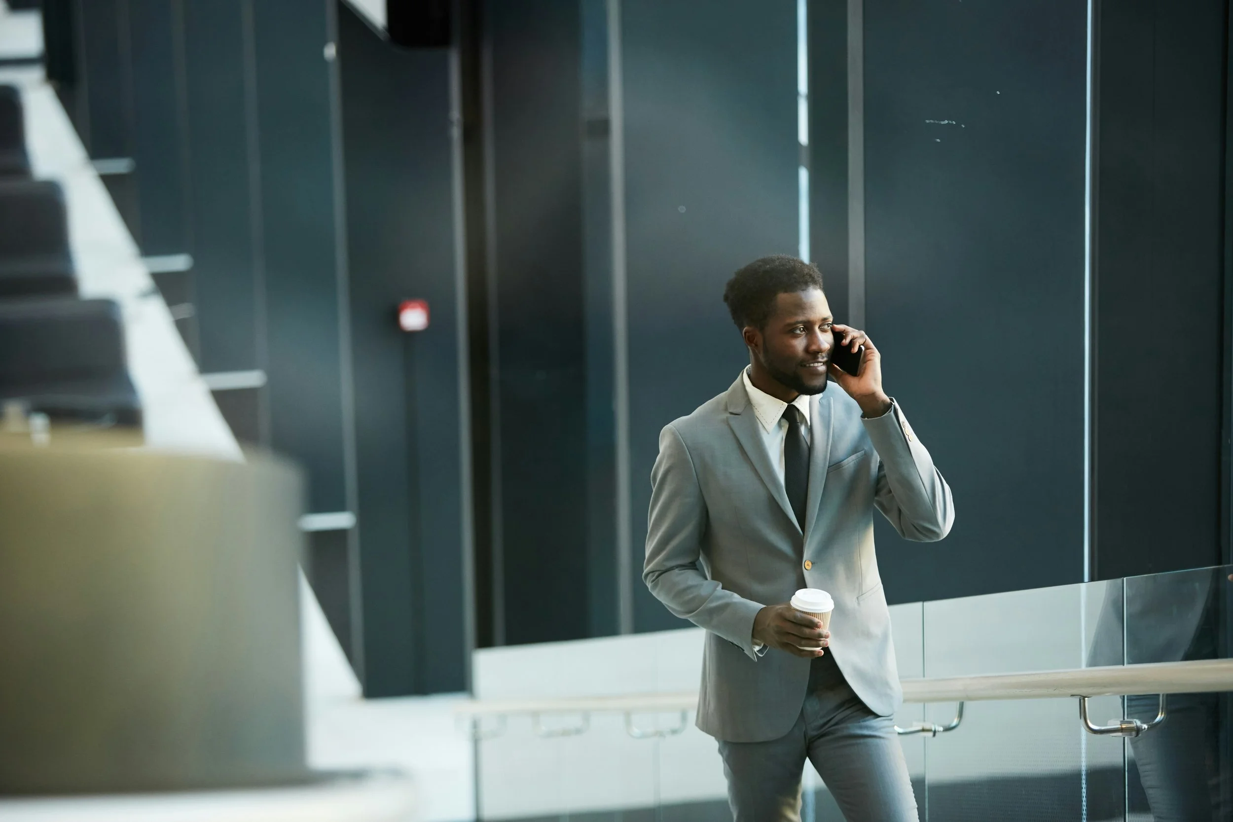 a business man holding his cellphone up to his ear, while holding a coffee, wearing a great business formal suit and a black tie