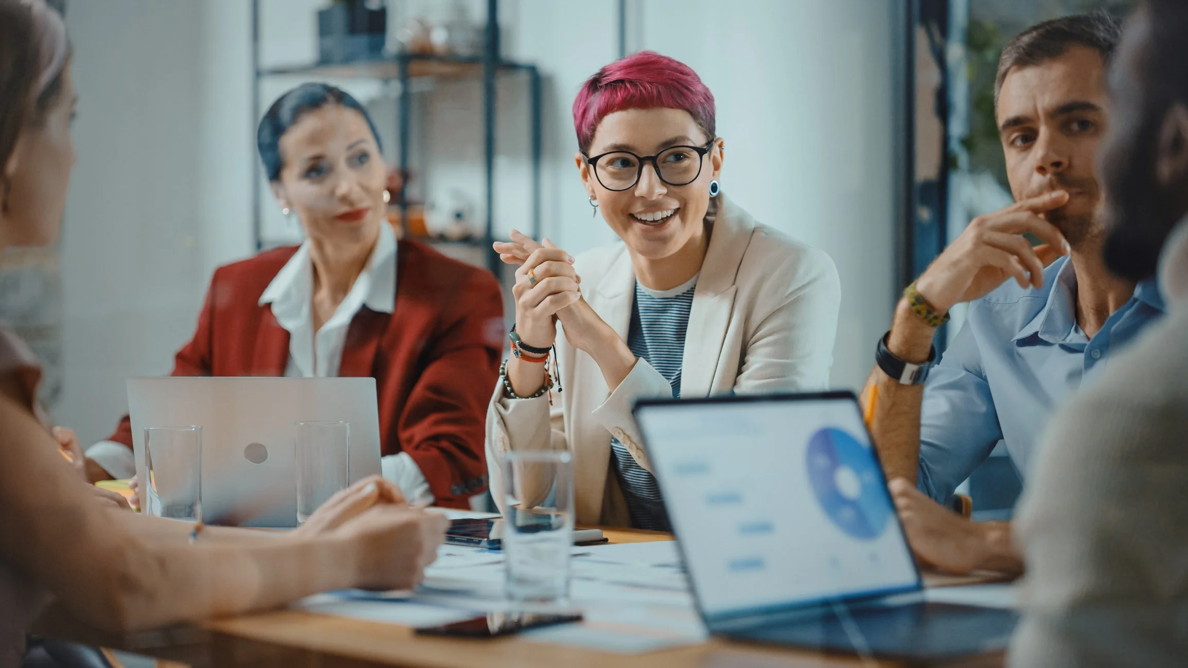 Group of people in a meeting room sitting around a table with laptops and documents, engaged in discussion. Nonbinary, neurodivergent person as center of focus.