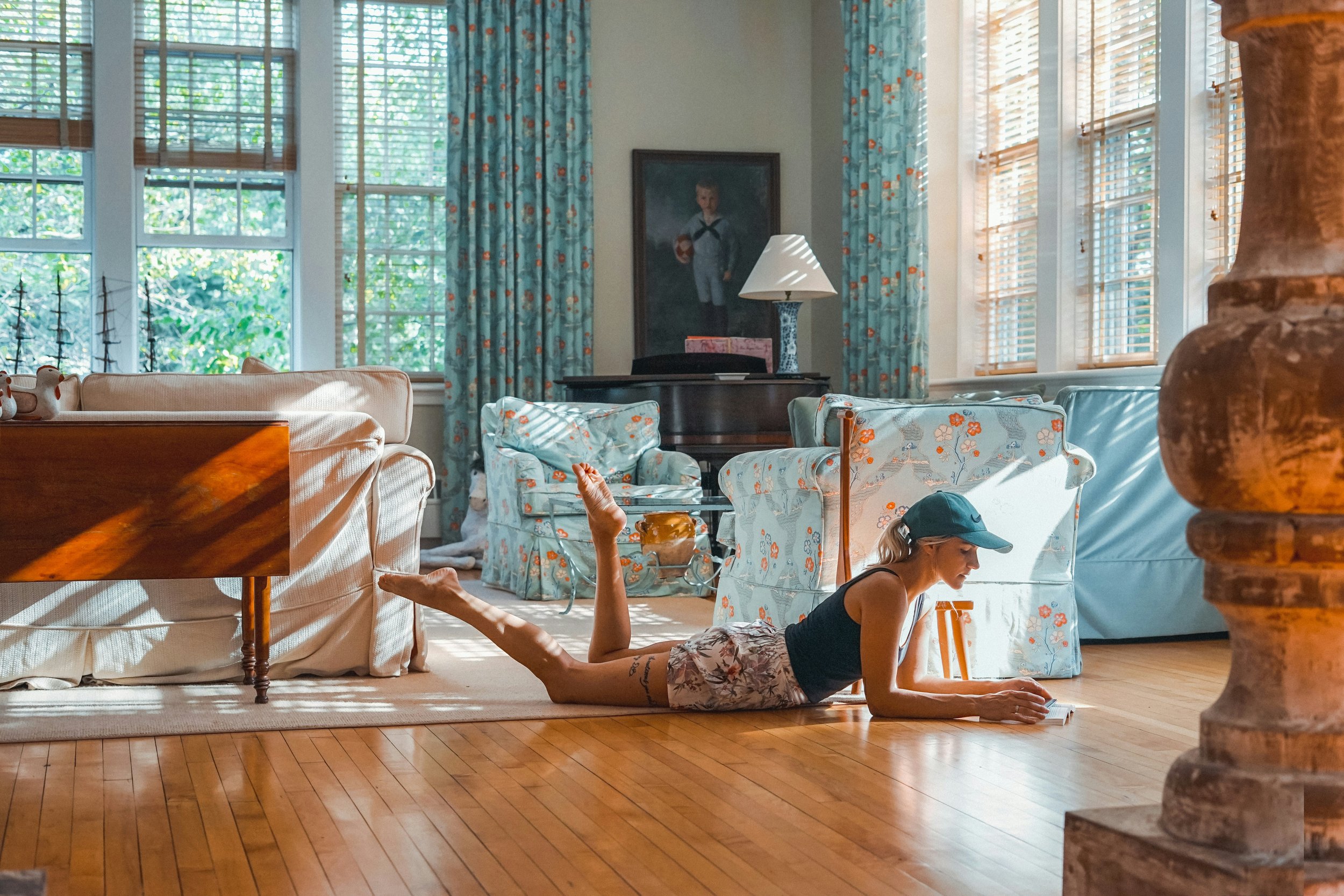 A woman in casual clothes, wearing a cap, lying on her stomach on the wooden floor of a living room, propped up on her elbows, reading a book.