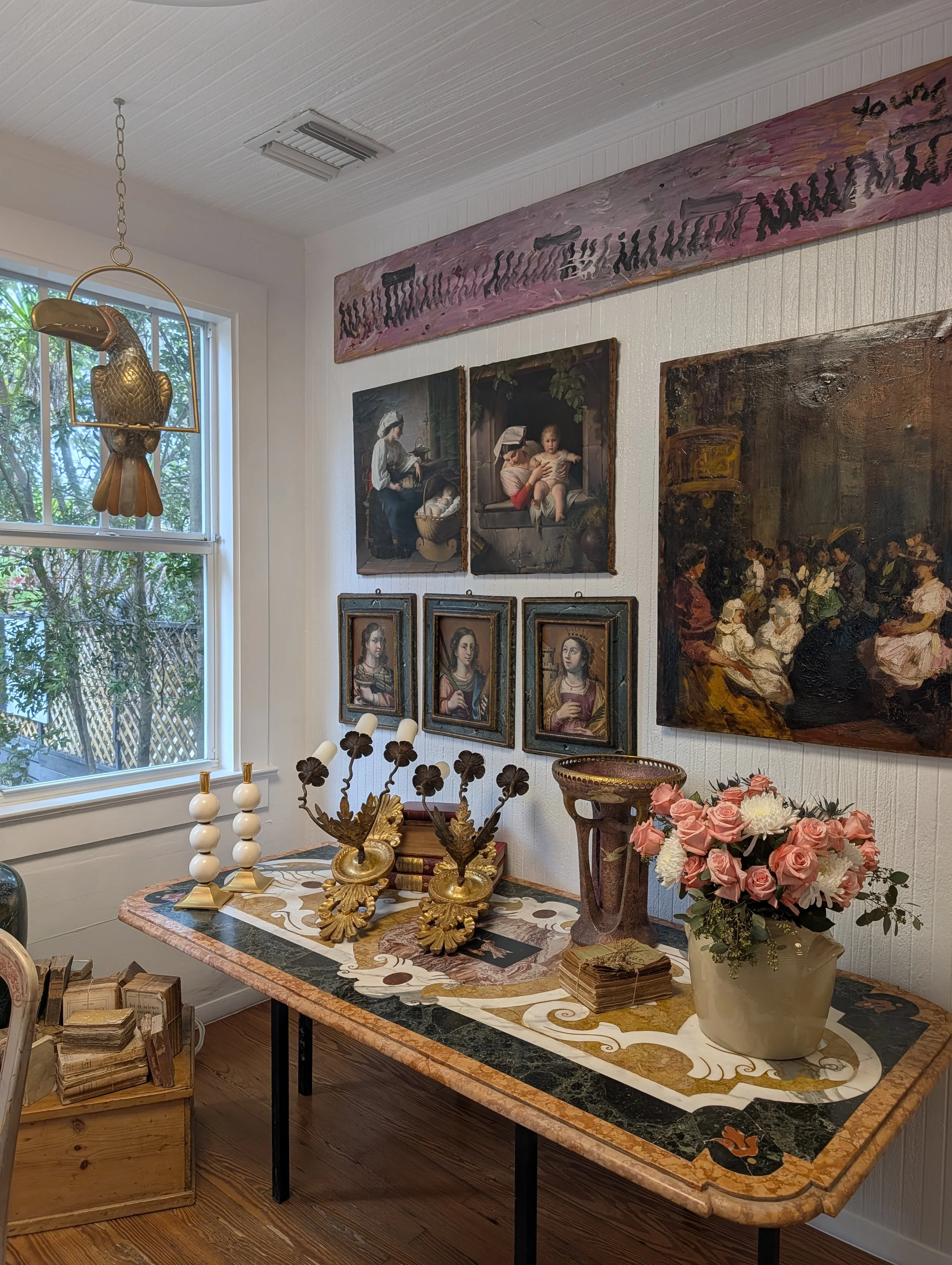 A room corner with a window, displaying framed classic oil paintings on the wall, atop an ornate table with decorative objects including a bouquet of pink and white flowers, candlesticks, a box, and a decorative urn.