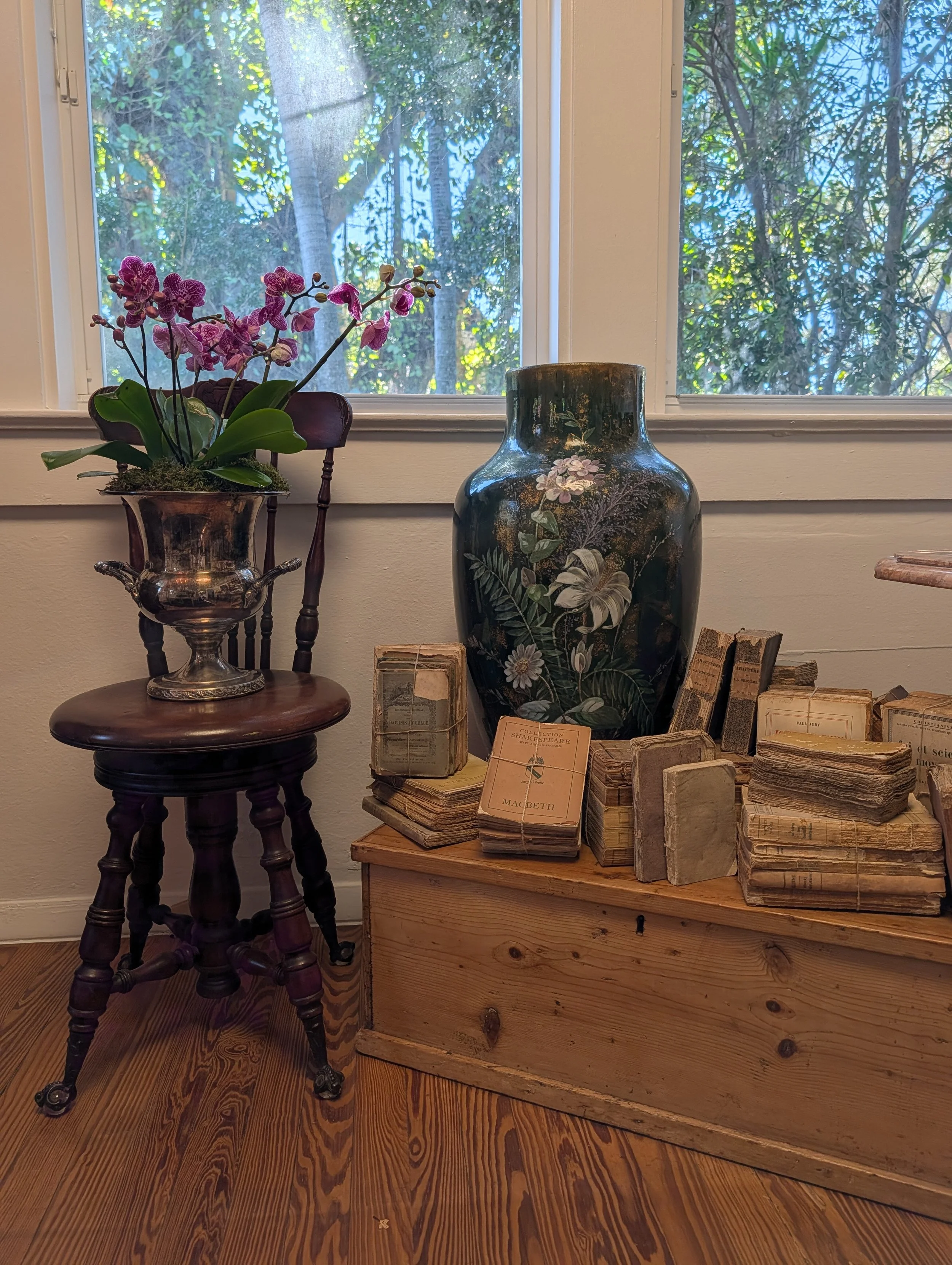 A wooden floor with a wooden box filled with old, worn books and papers, a large decorative vase with floral paintings, a silver pot holding a potted orchid, and a wooden chair behind the orchid. A window with trees outside is in the background.