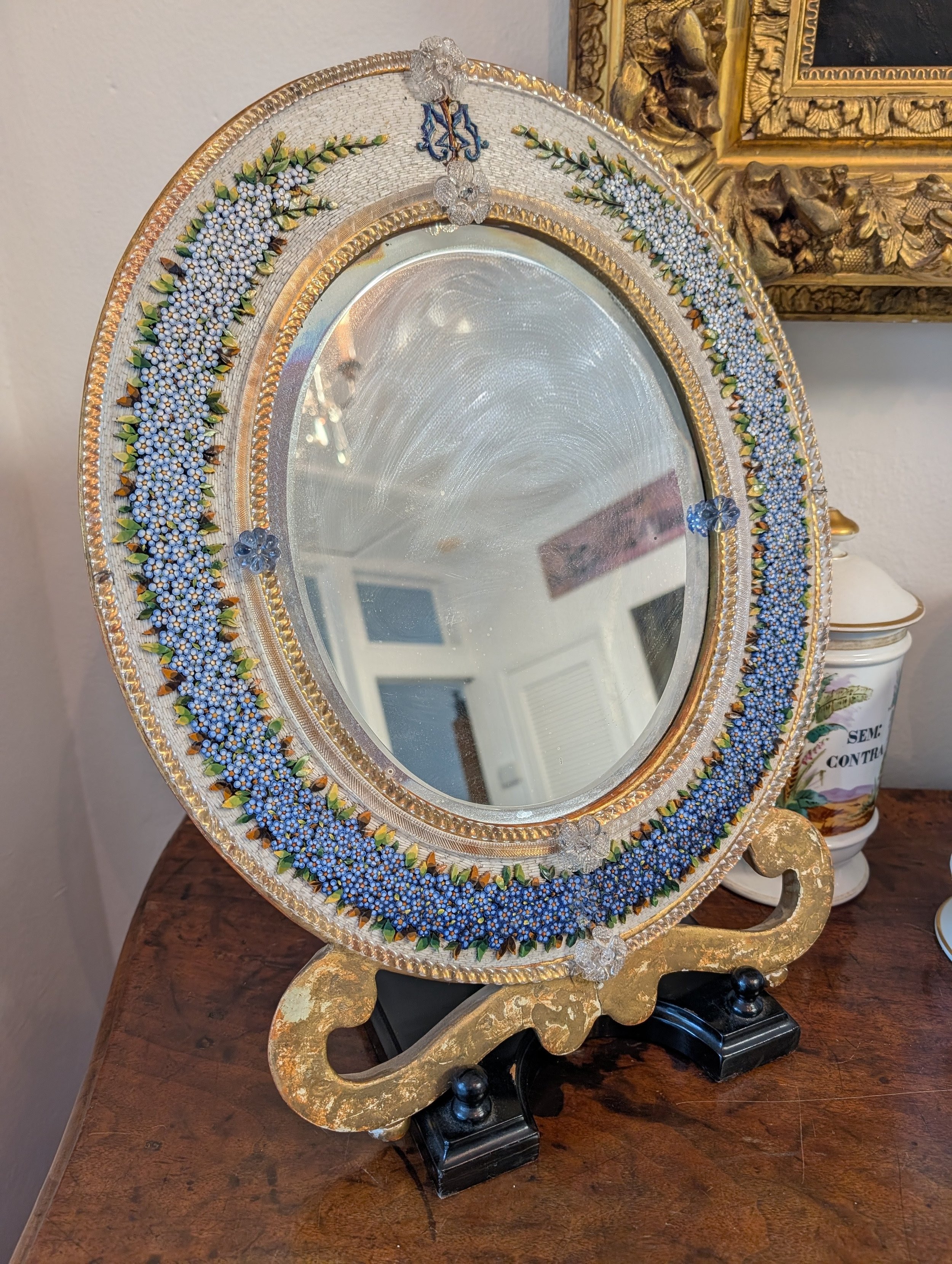 Decorative oval mirror with a micromosaic floral border, set on a gold and black stand, placed on a wooden surface.