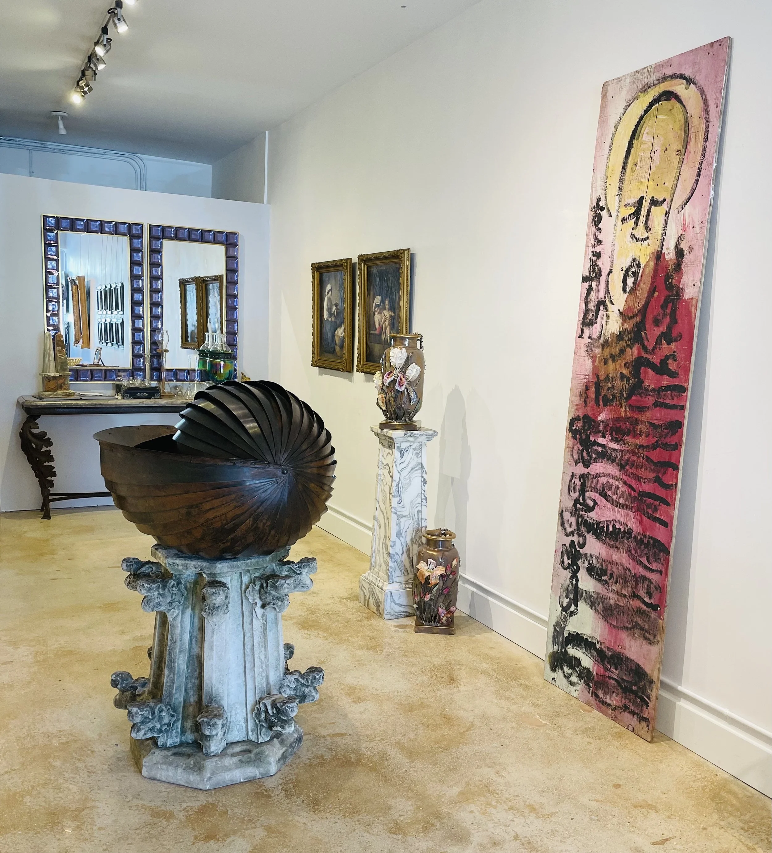 Art gallery with sculptures, paintings, and mirrors, including a large dark brown spiral sculpture on a carved stone pedestal, smaller vases with floral designs, and framed paintings on a white wall.