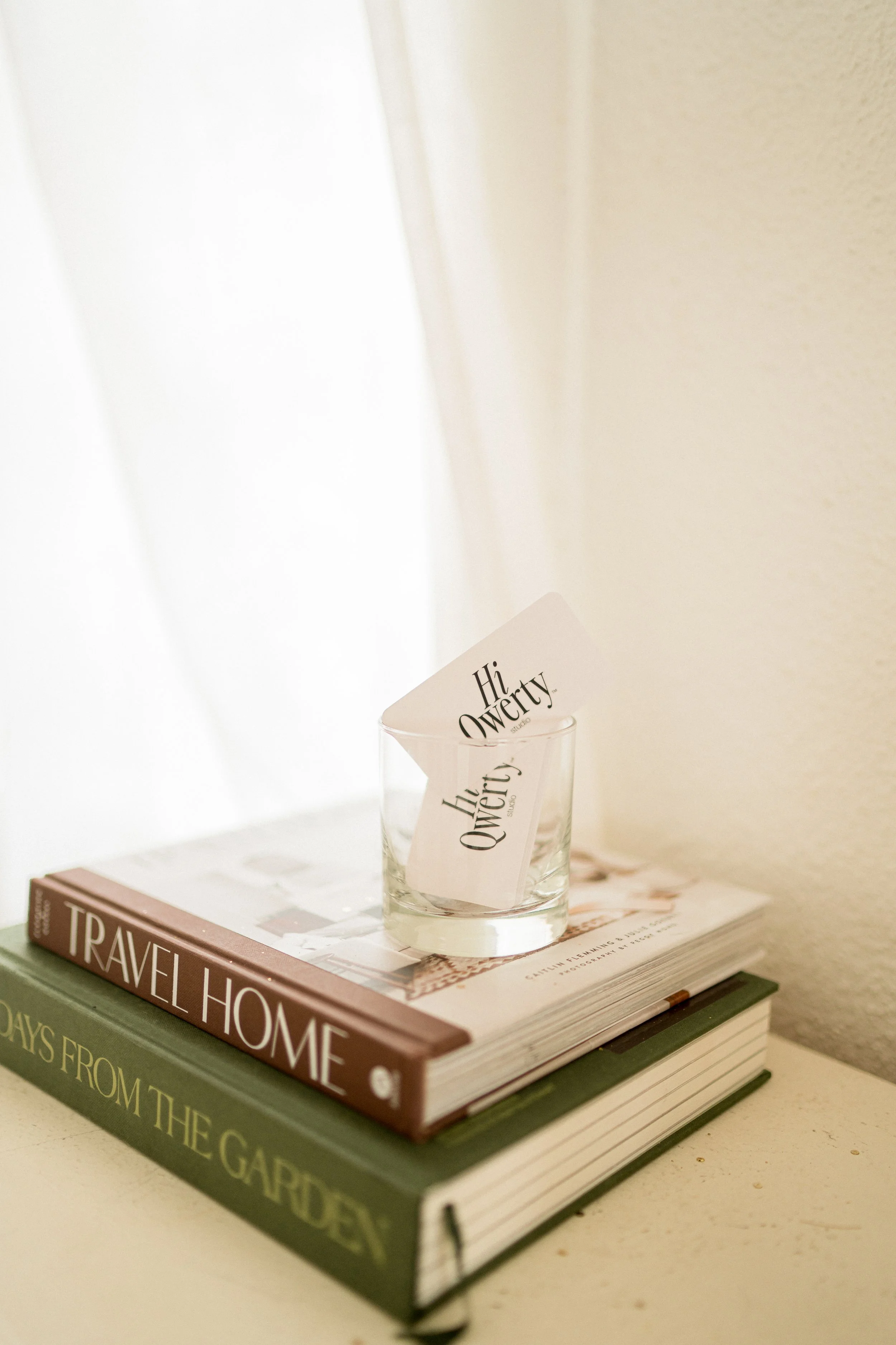 A clear glass with a 'Hi, Querty' note inside, placed on top of two stacked books titled 'Travel Home' and 'Days From the Garden' on a white surface next to a white wall.