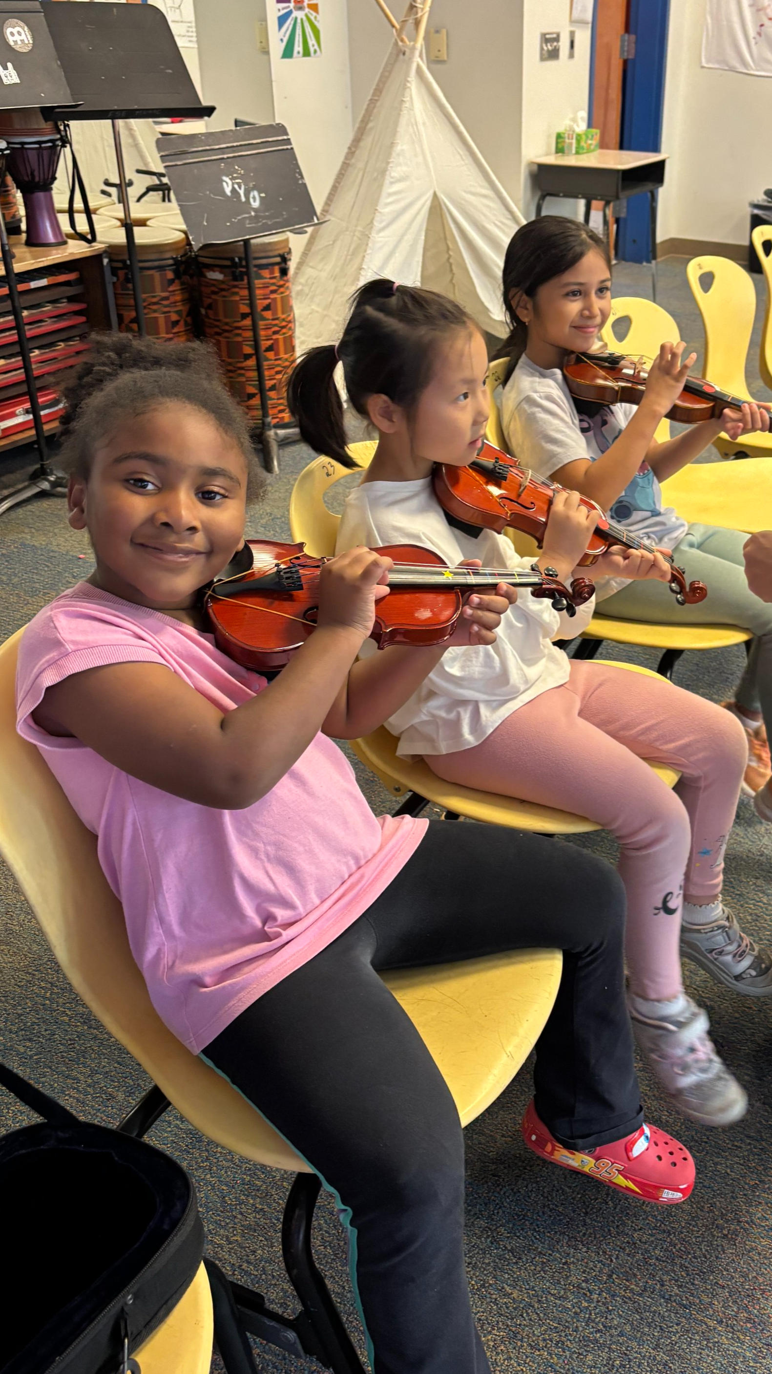 Three young girls sitting in yellow chairs, playing violins during a music lesson.