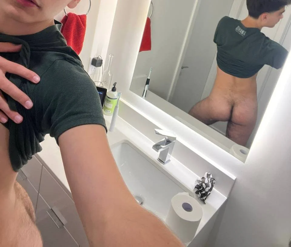 A person taking a selfie in a bathroom mirror, showing their back and partly revealing their face. The bathroom has a white sink counter, a roll of toilet paper, a hand soap dispenser, and a red towel hanging on the wall. The person's reflection in the mirror shows them without pants, and they are wearing a dark shirt. The photo captures the bathroom and the person's partial nudity.