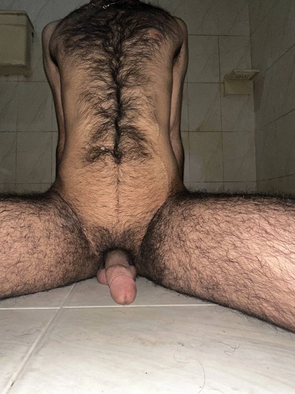 A naked man sitting on the floor, seen from below, with his legs spread apart.