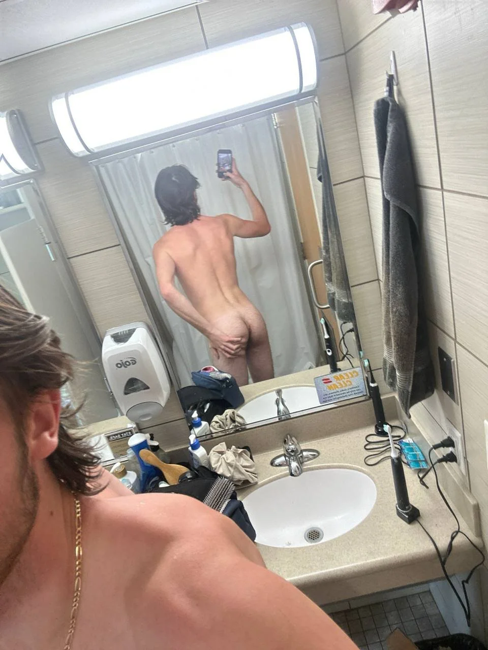 A man taking a mirror selfie in a bathroom, with a second man visible from behind in the mirror, both appear to be naked.