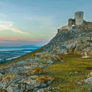 A Romanian castle perched on a hilltop overlooking a landscape with water and distant landforms during sunset.