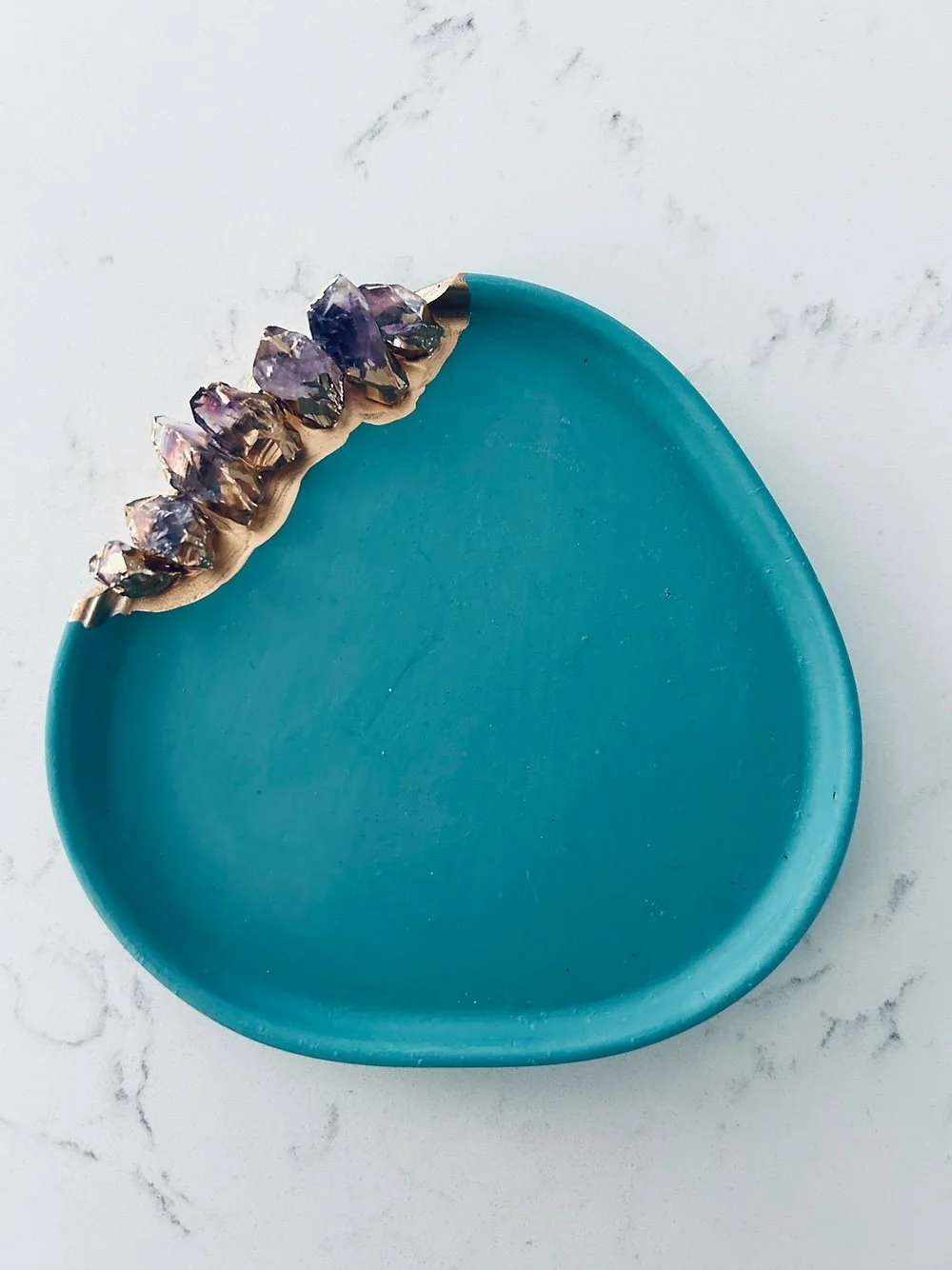 A teal decorative dish with embedded crystals along the edge, placed on a marble surface.