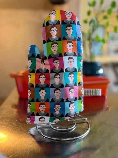 A colorful oven mitt featuring a pop art-style pattern of a person wearing glasses and a collar, repeating in different color backgrounds, displayed on a metal stand in a kitchen setting.