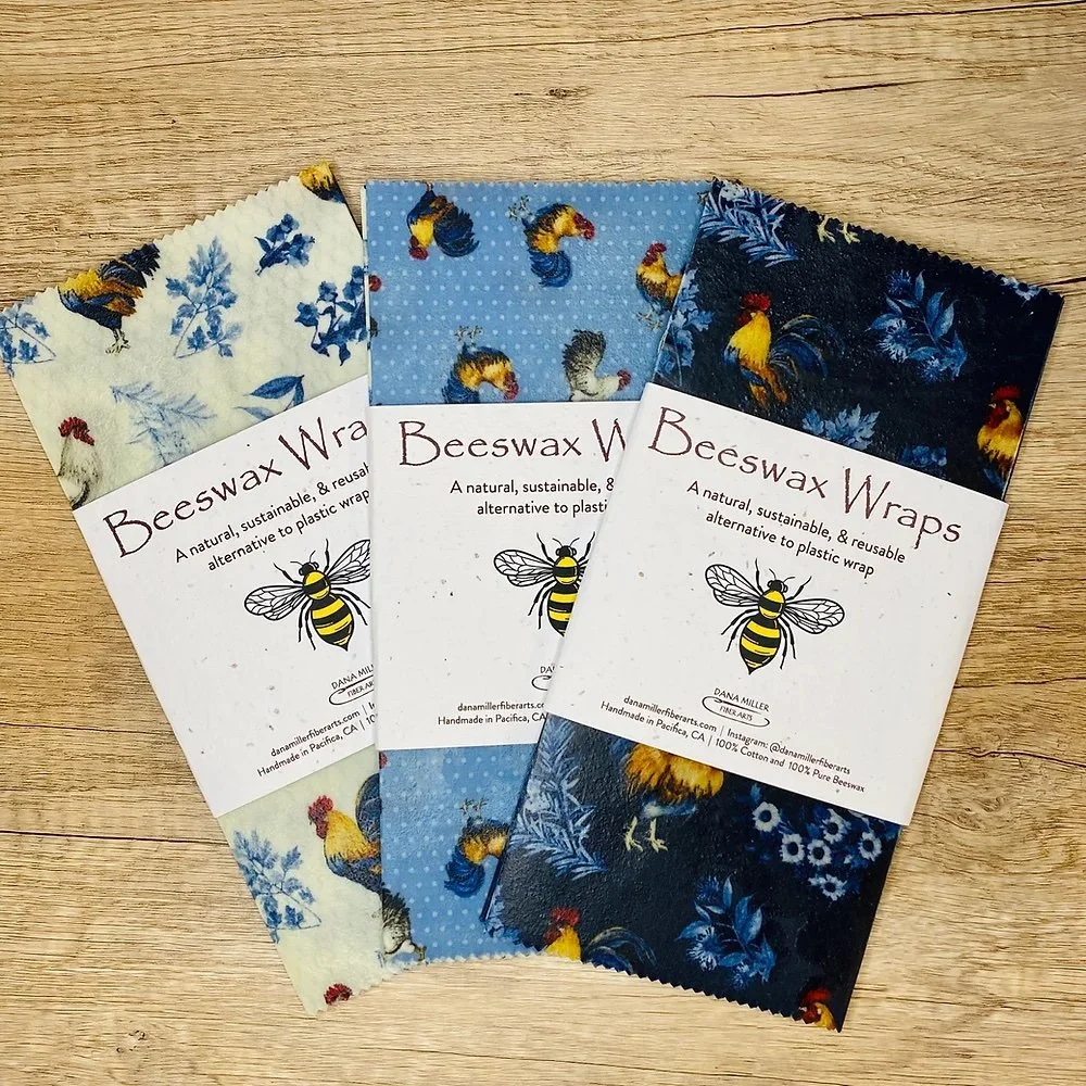 Three packs of beeswax wraps with rooster and floral patterns on a wooden surface.