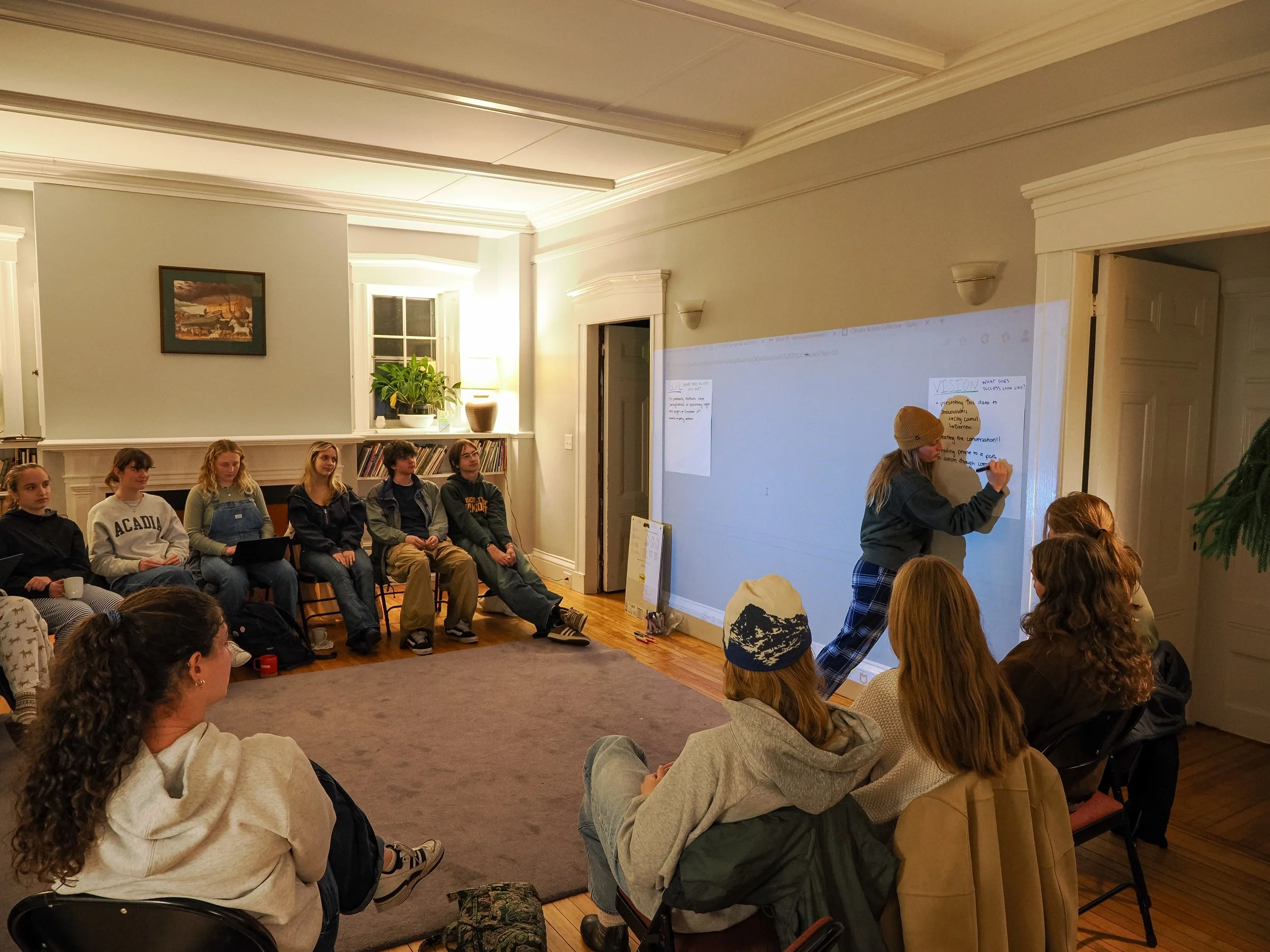 College students gathered in a circle watching a presentation while the facilitator writes on the board
