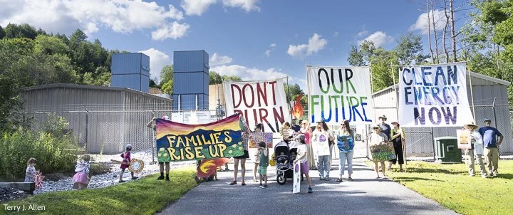 A group of adults and children stand in front of a fenced industrial compound holding banners that say "Families Rise Up," "Don't Burn Our Future," and "Clean Energy Now"