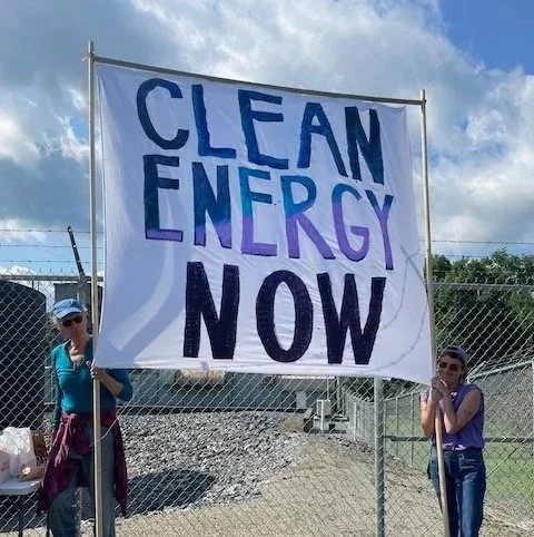 Activist hold a sign saying "Clean Energy Now" in front of the fenced in Ascutney, VT Peaker Plant