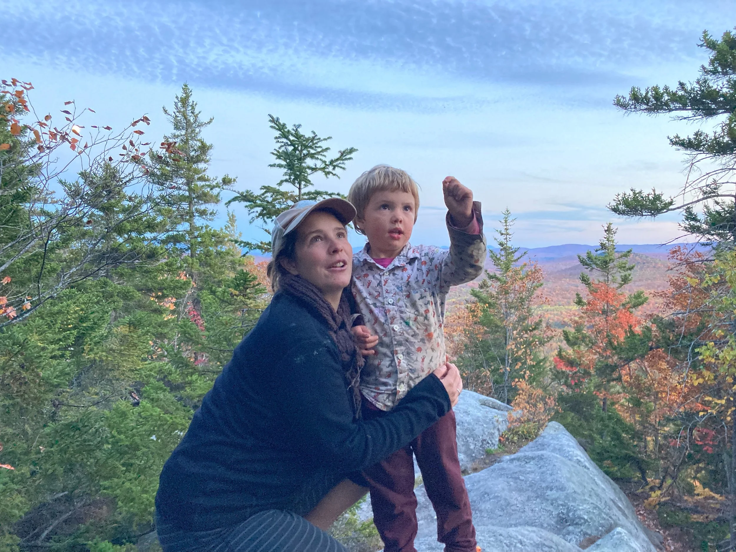 A person in a baseball hat crouching next to a child in front of trees and a mountain view.
