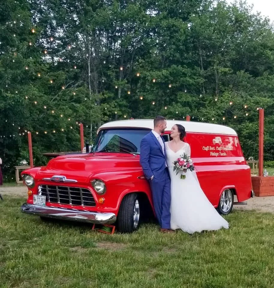 A bride and groom standing together, holding hands and looking at each other, beside a vintage red mobile bar with a sign that reads 'Craft Beer, Craft Cocktails, Vintage Trucks,' in a grassy outdoor setting with string lights in the background.