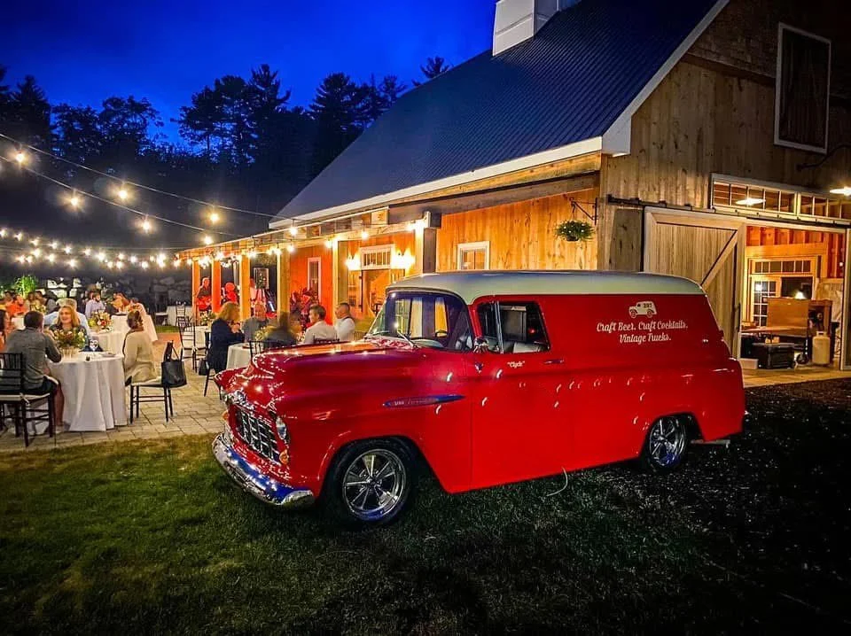 Outdoor wedding reception at dusk with guests sitting at round tables with white tablecloths, string lights overhead, a rustic wooden barn, and a red vintage Chevrolet mobile bar parked on the grass.