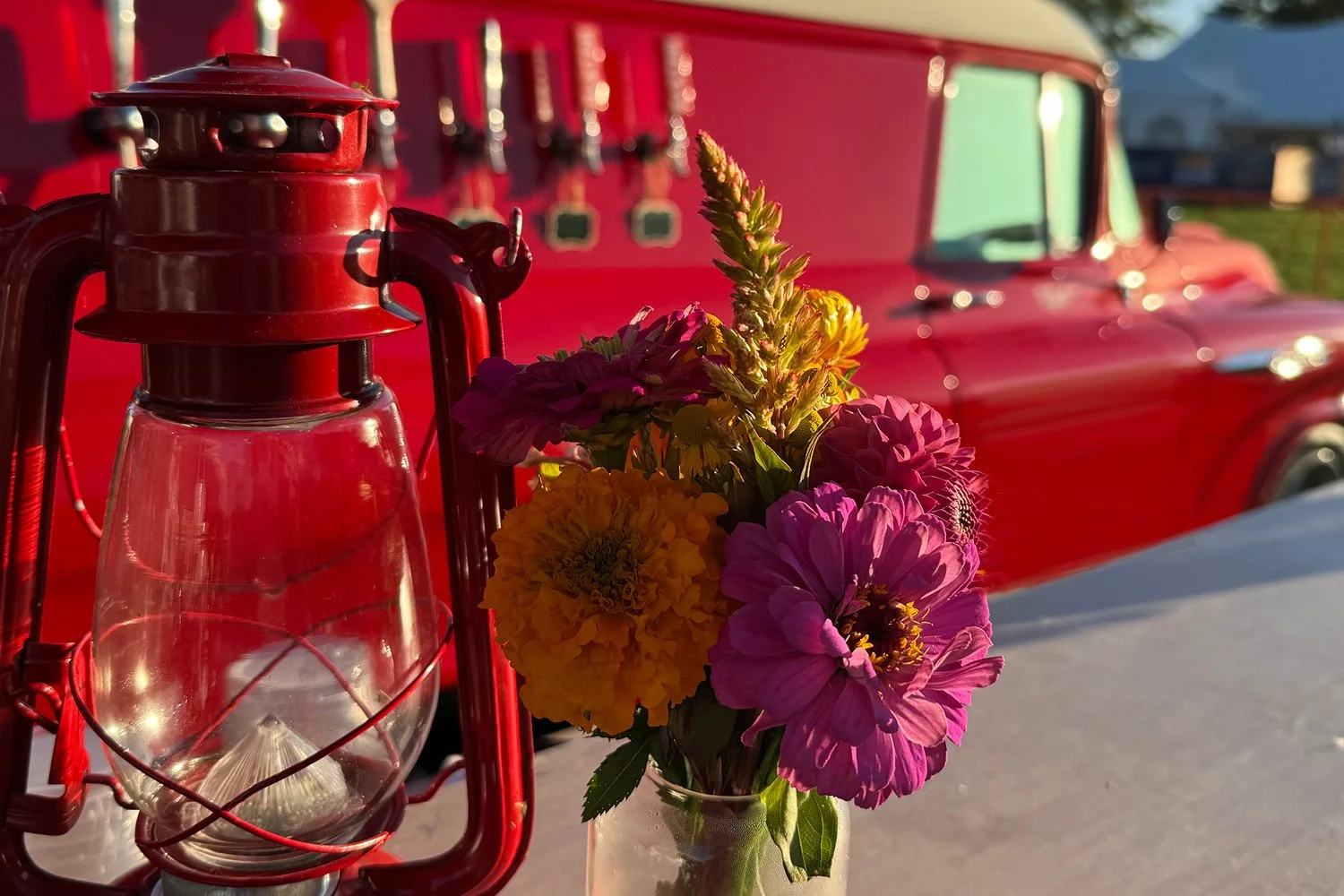 A red vintage lantern and a small bouquet of colorful flowers in a glass jar, with a red vehicle in the background