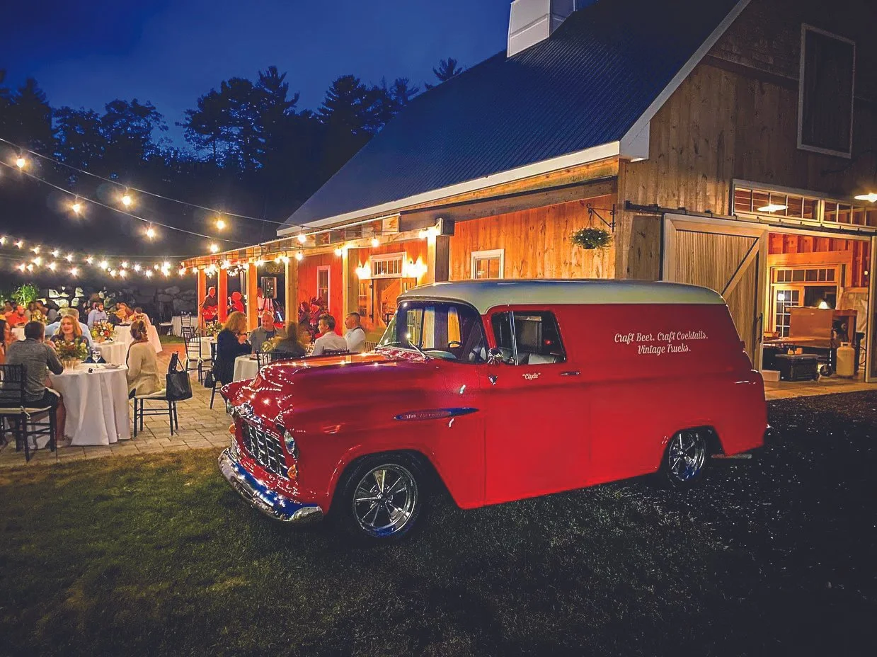 Red vintage Chevrolet mobile bar parked outside a rustic barn-style building with outdoor dining and string lights at night.